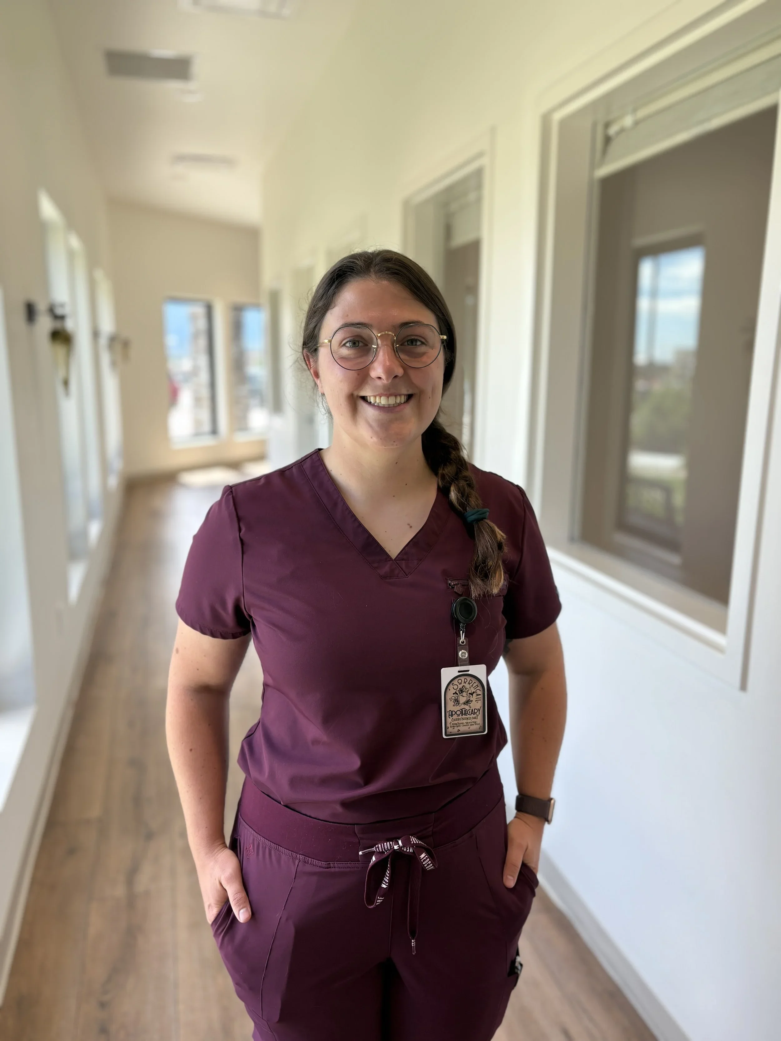 A woman with glasses and a braid, wearing scrubs, standing in a brightly lit corridor with windows, smiling at the camera.