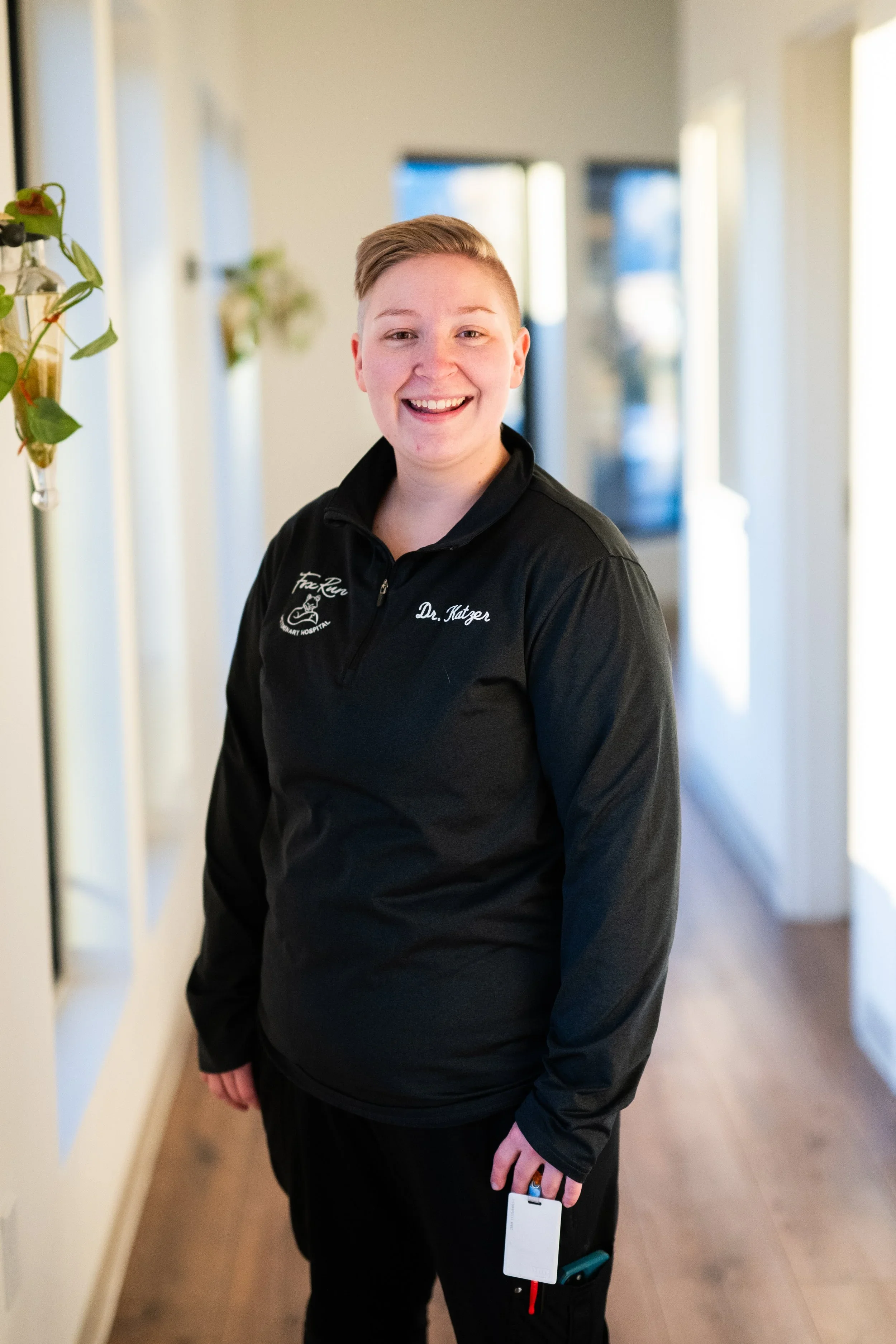 A smiling woman with short hair wearing a black jacket with a logo and name 'Dr. Katzer' standing in a hallway with white walls and wooden flooring, holding a white card and a pen.