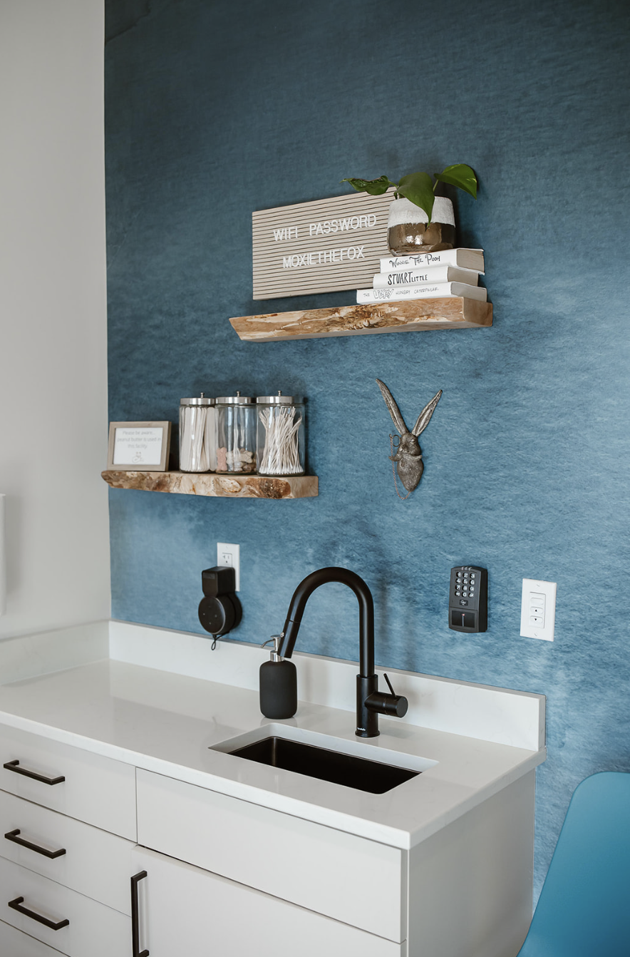 Modern kitchen with white cabinets, a white countertop, a black faucet over a black sink, blue textured wall, wooden shelves with decorative items, jars with cotton swabs and pink items, a rabbit wall decoration, and electrical outlets with a phone a