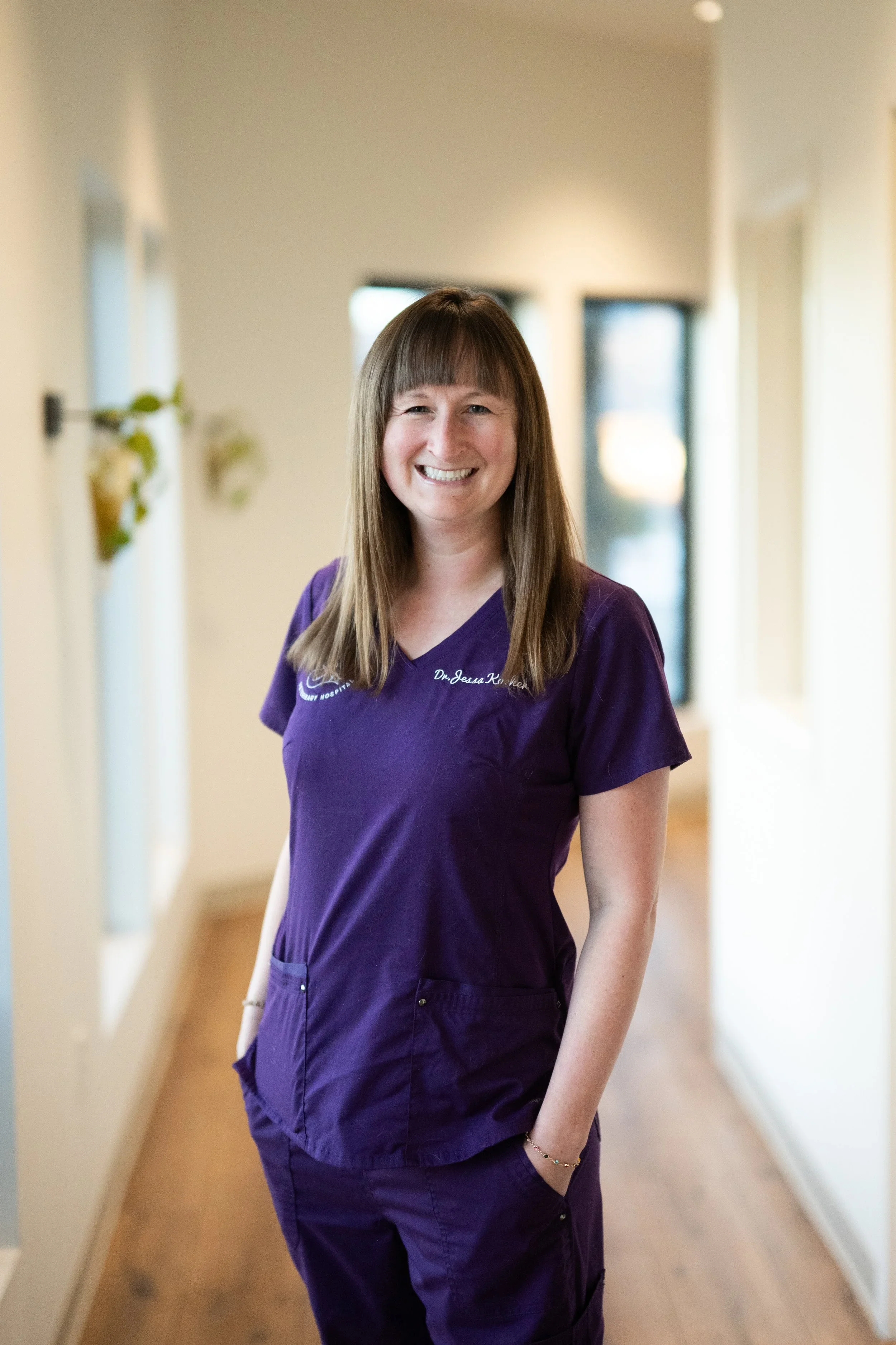 A woman with long brown hair wearing purple scrubs standing in a hallway with light-colored walls and wooden flooring, smiling at the camera.