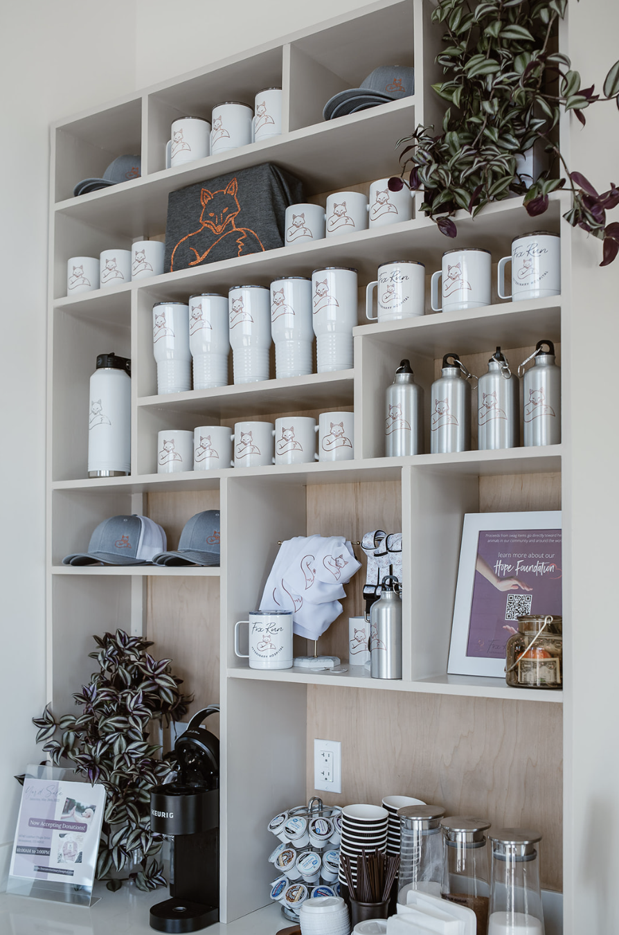 A display shelf with white coffee mugs, water bottles, caps, and a framed sign, decorated with a plant.