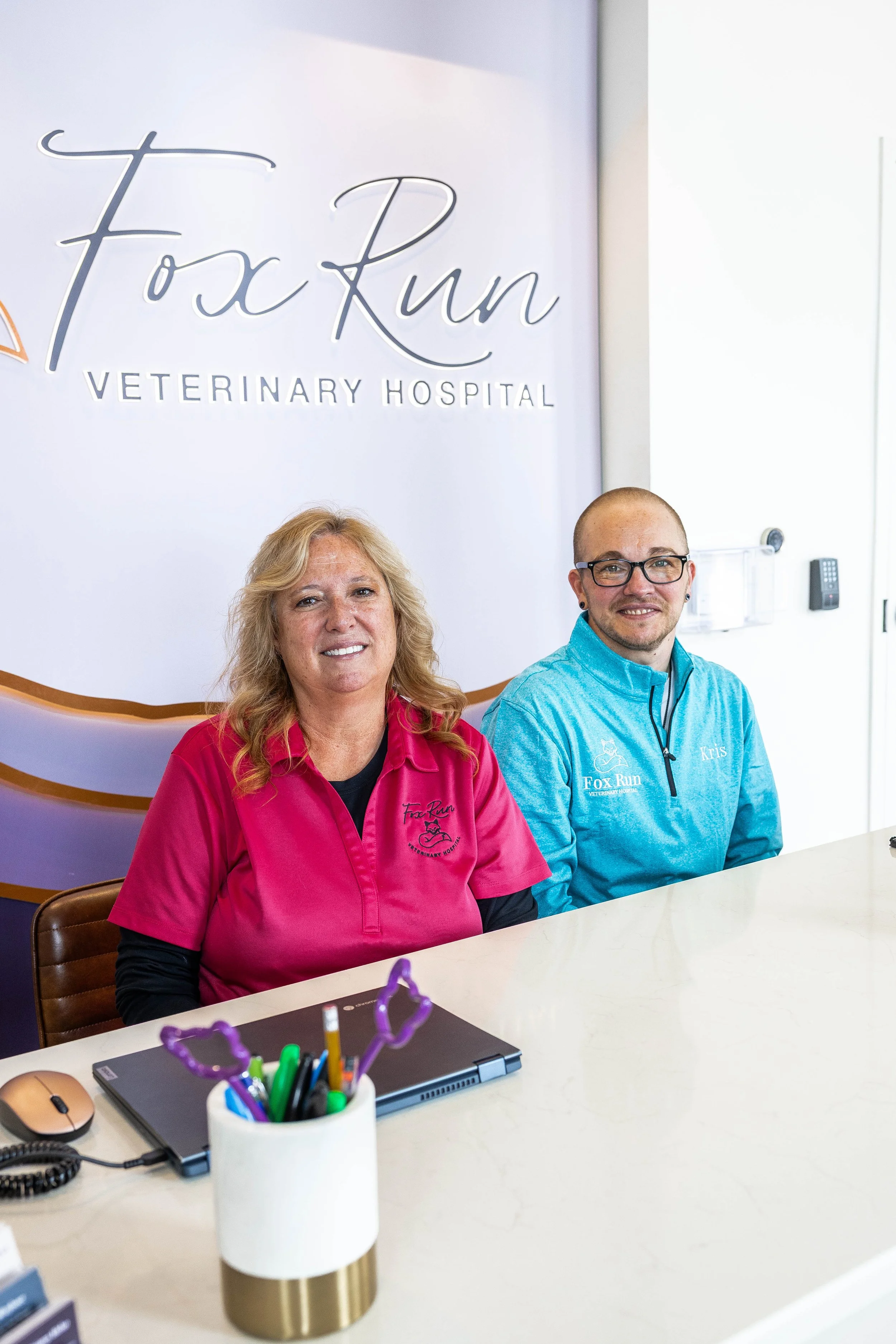 Two veterinary professionals sitting at a reception desk in a veterinary hospital. The woman wears a pink uniform, and the man wears a blue uniform with a name tag. Behind them, there is a sign that reads "Fox Run Veterinary Hospital." On the desk, there is a laptop, a telephone, and a pen holder with various pens and scissors.