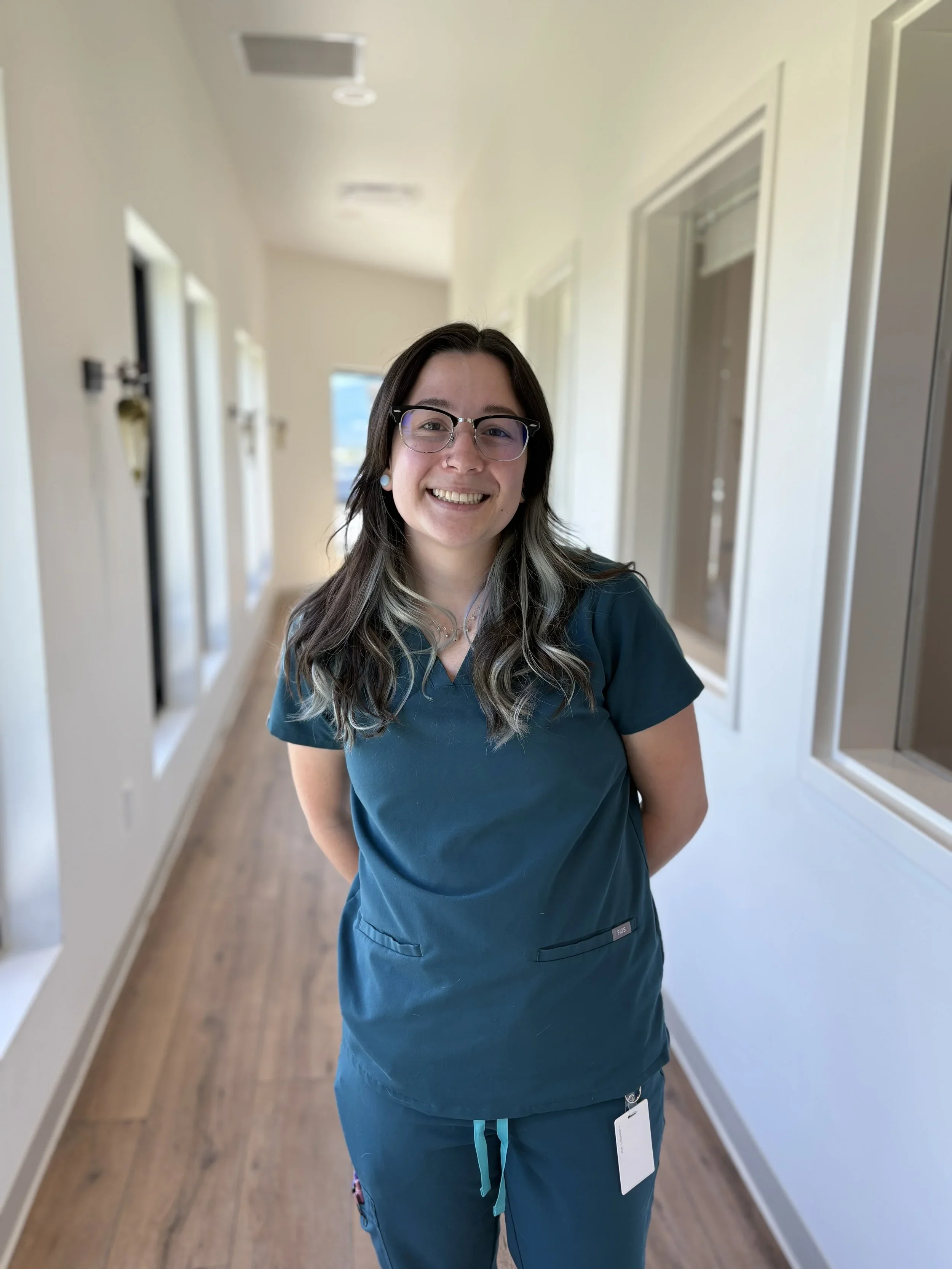 A young woman wearing blue medical scrubs, glasses, and earrings, smiling, standing in a well-lit hallway with light-colored walls and wooden flooring.