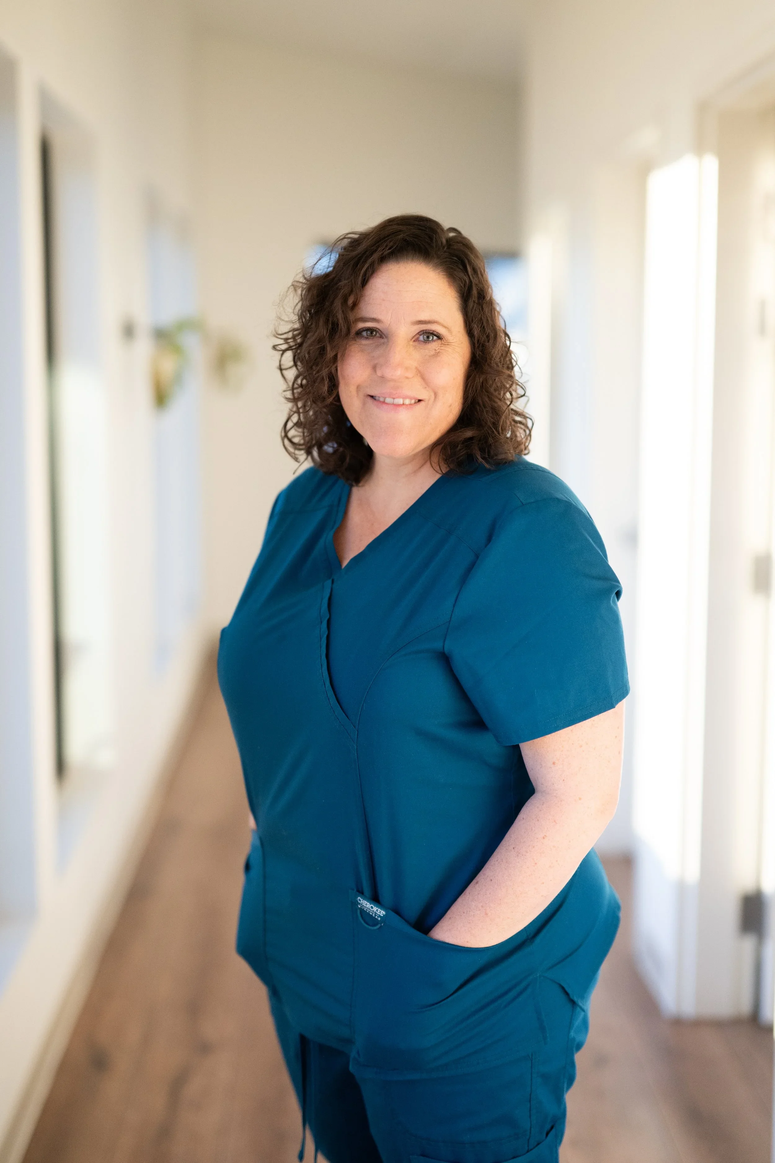 A woman with curly brown hair wearing blue medical scrubs standing in a well-lit hallway, smiling at the camera.
