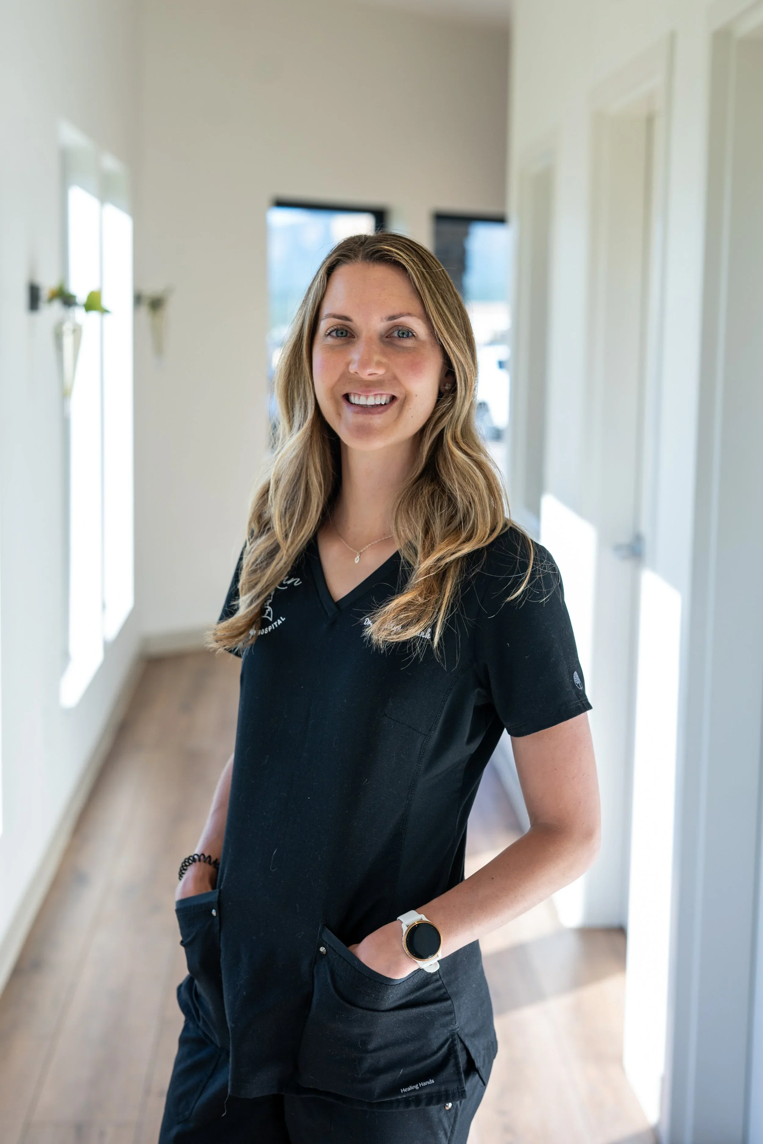 A young woman with long, wavy blonde hair smiling and wearing a black medical uniform with her hands in her pockets, standing in a bright, modern hallway with white walls and wooden flooring.