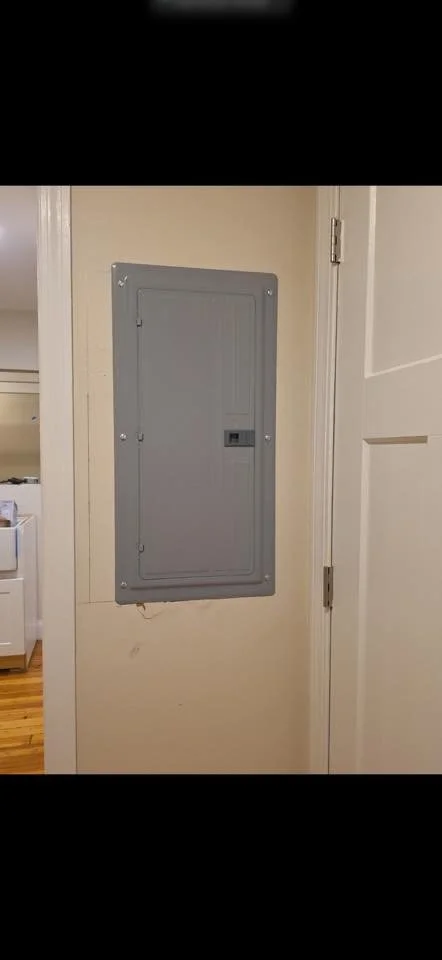 A gray electrical panel mounted on a beige wall next to a white door in a room with hardwood floors.
