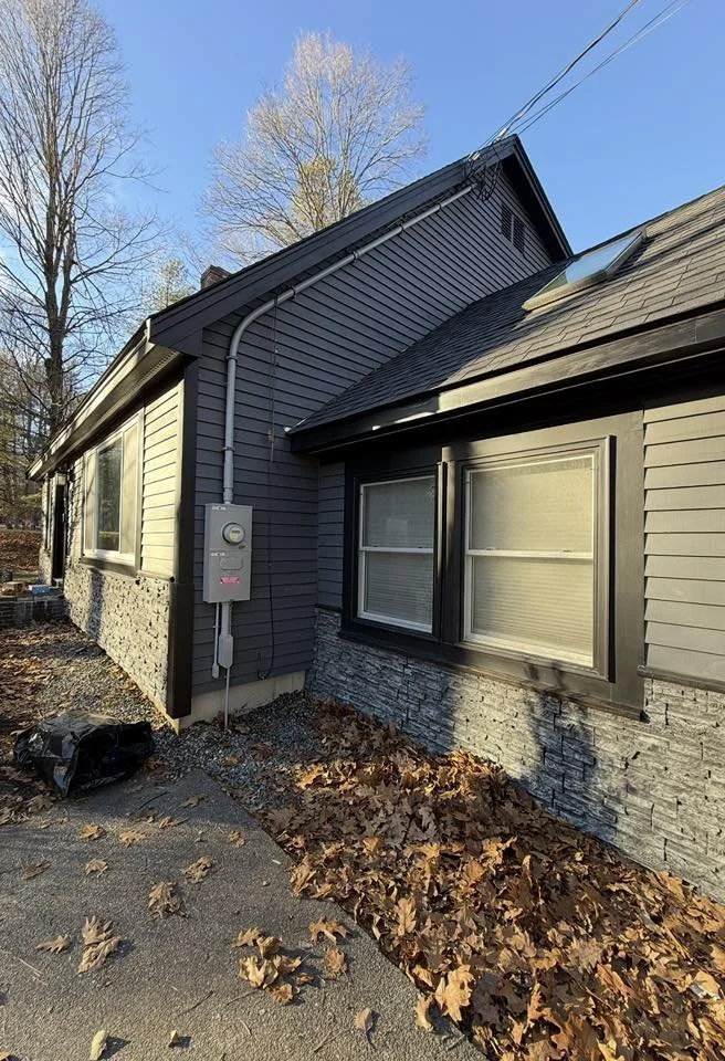 A house exterior with gray siding and stone foundation, two large windows, a skylight, fallen leaves on the ground, and a utility meter on the wall.