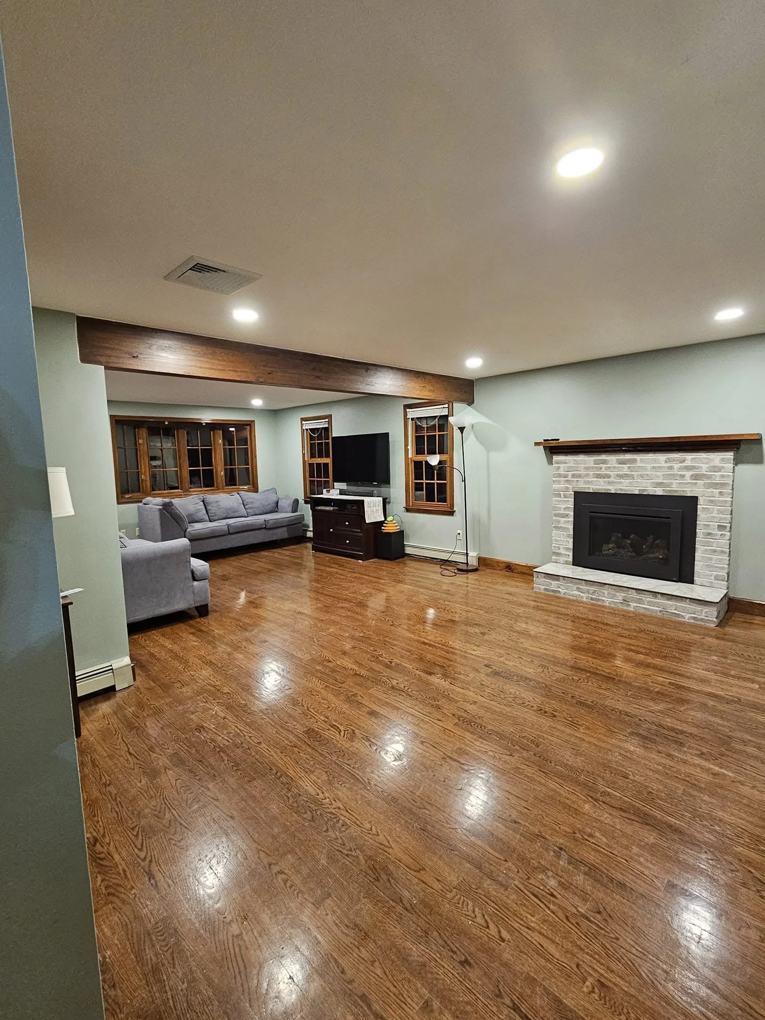 Living room with hardwood floors, light green walls, a brick fireplace, multiple ceiling lights, a gray sofa, a television, and windows with wooden frames.