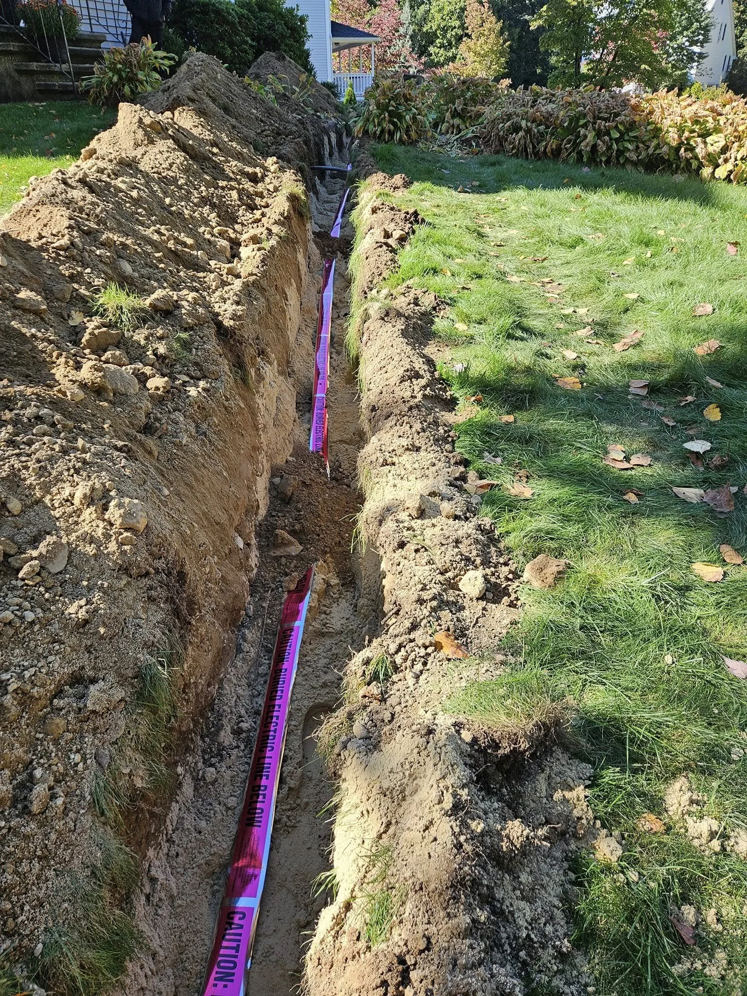 A narrow trench dug in a person's front yard, with purple caution tape along its length indicating underground utility lines. The trench is adjacent to a grassy lawn with fallen leaves, in a suburban neighborhood with trees and houses visible in the 