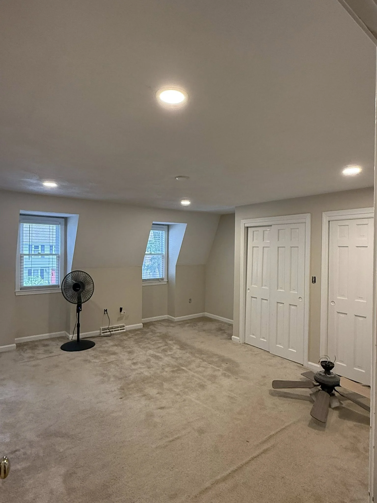 A spacious room with beige carpet, three windows with blinds, two white closet doors, a black oscillating fan, and some scattered furniture pieces, including a fan and a small table.