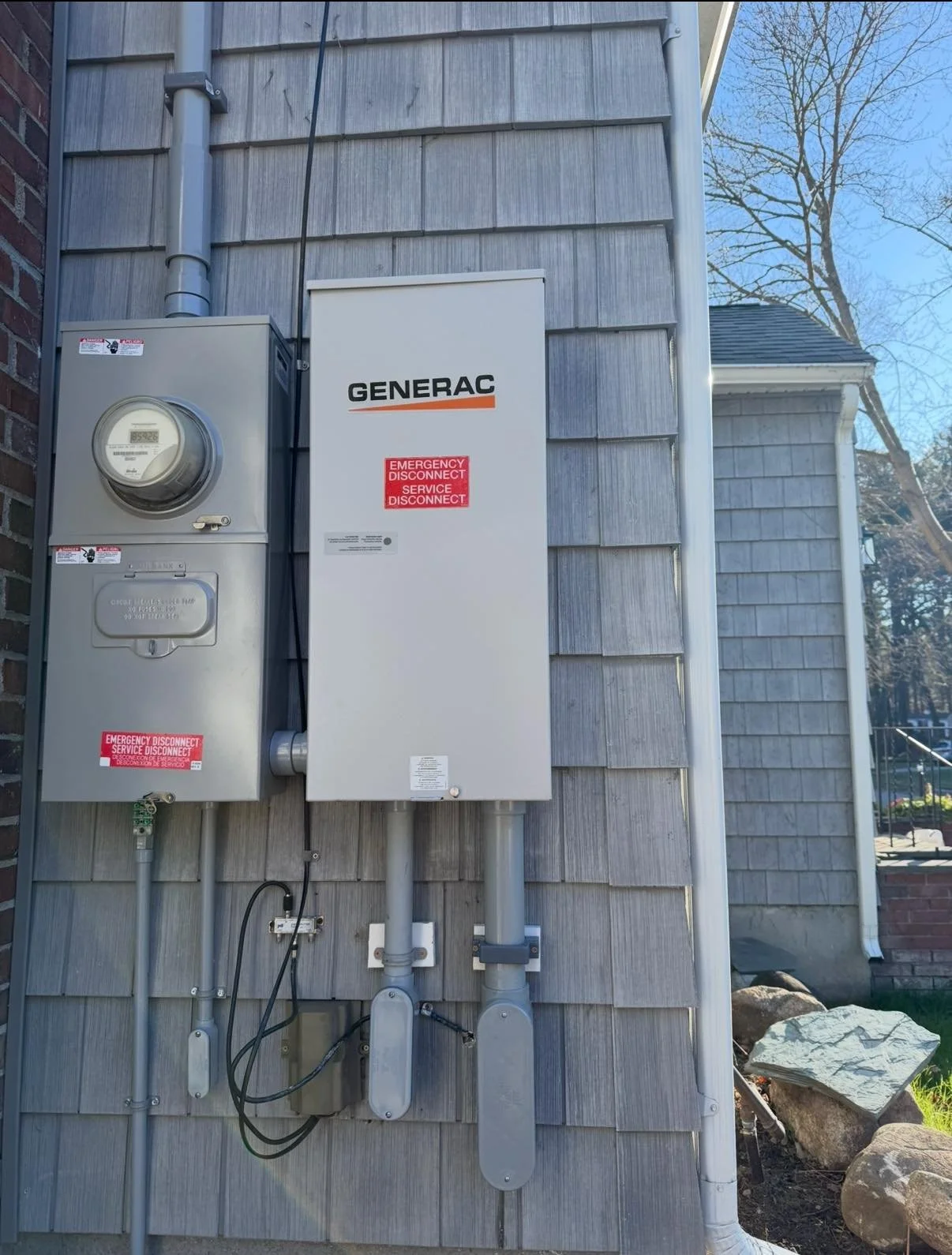 Generator setup on the exterior wall of a house with electrical boxes and piping, gray siding, some trees in the background, and a blue sky.