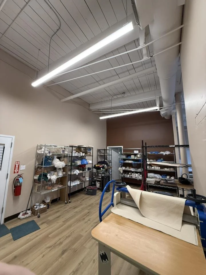 A storage room with metal shelving units filled with various supplies. There are fluorescent lights on the ceiling, an industrial paper folding machine on a wooden table in the foreground, and a fire extinguisher mounted on the wall to the left.