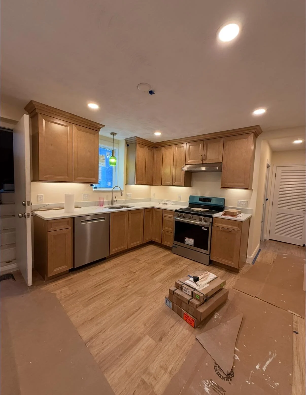 Kitchen with wooden cabinets, stainless steel appliances, a white countertop, a small window, and a pendant light. There are construction boxes on the floor, indicating ongoing renovation.