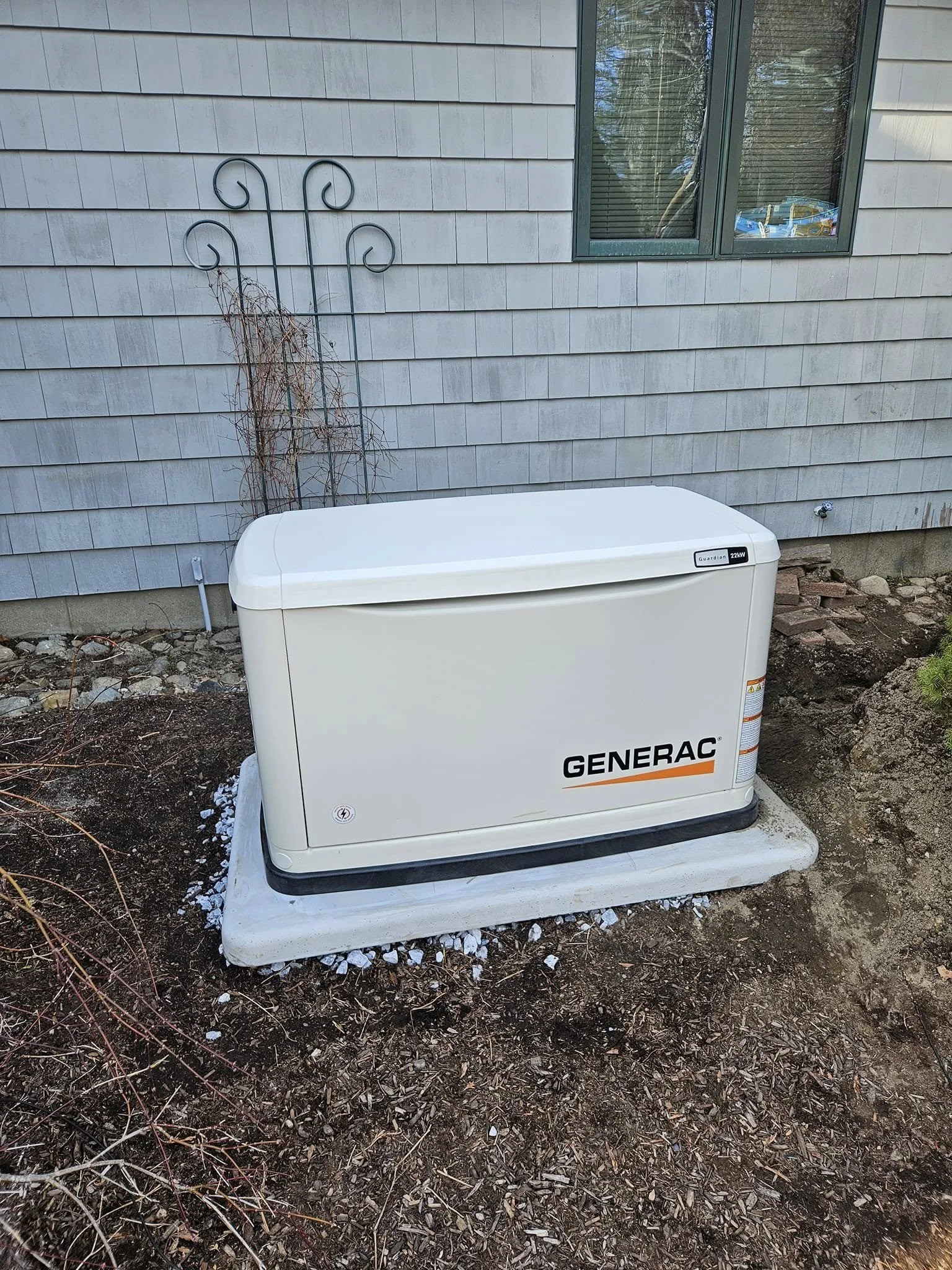 A Generac Guardian 22kW home standby generator installed outside a house with gray siding, next to a window, on a concrete pad surrounded by dirt and wood chips.