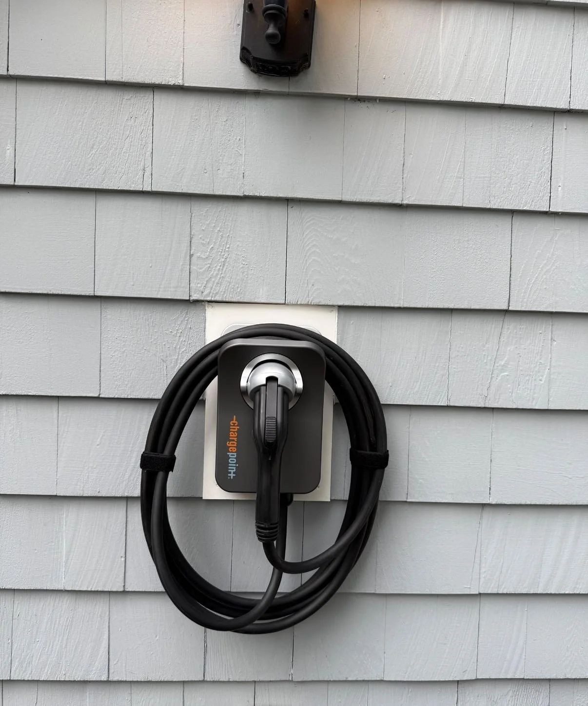 An electric vehicle charging station mounted on a gray wooden exterior wall, with a black charging cable coiled around it and a black power outlet above.