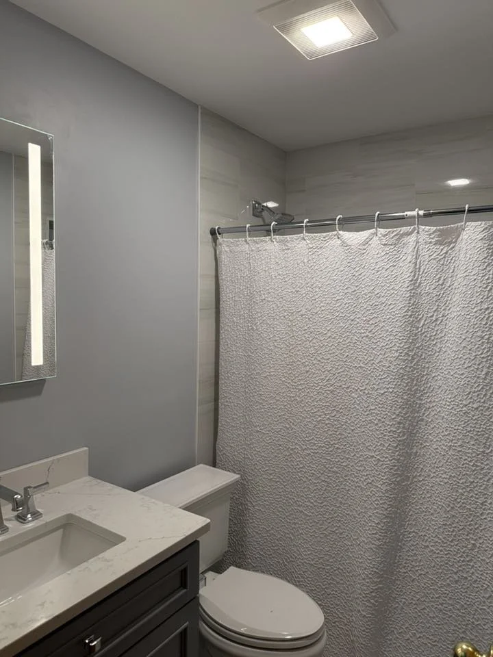Bathroom with light gray walls, a mirror, a white marble countertop sink, a dark gray vanity, a toilet, and a shower with a textured white shower curtain.