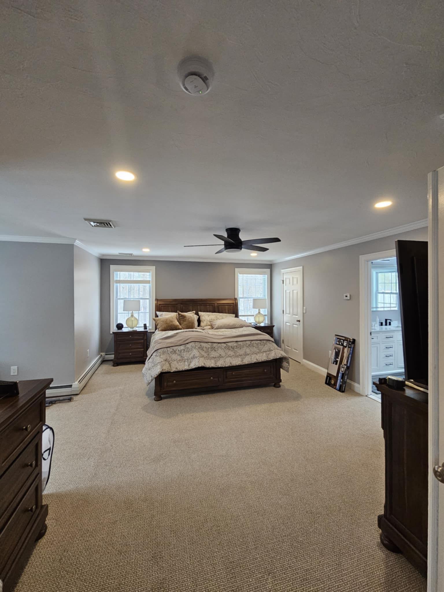 A bedroom with a bed in the center, two nightstands with lamps, windows behind the bed, ceiling fan, and beige carpet.