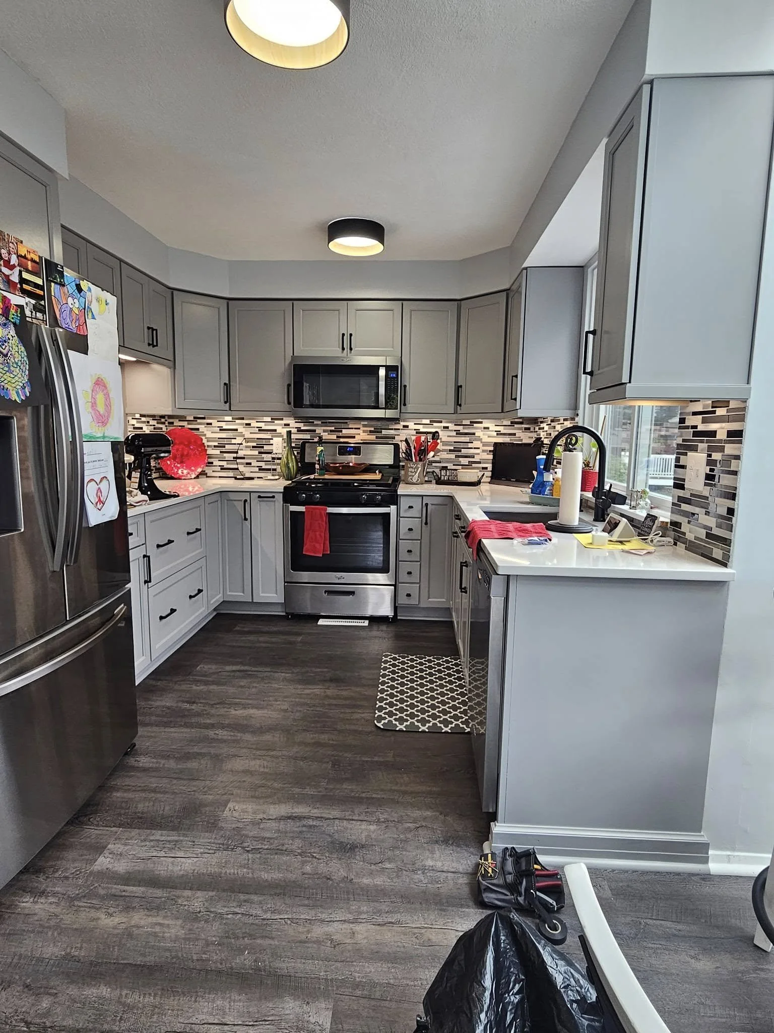 A modern kitchen with gray cabinets, stainless steel appliances, a black and white tile backsplash, and a window. Items on the counter include a paper towel roll, a soap dispenser, and various tools, indicating ongoing work or cleanup.