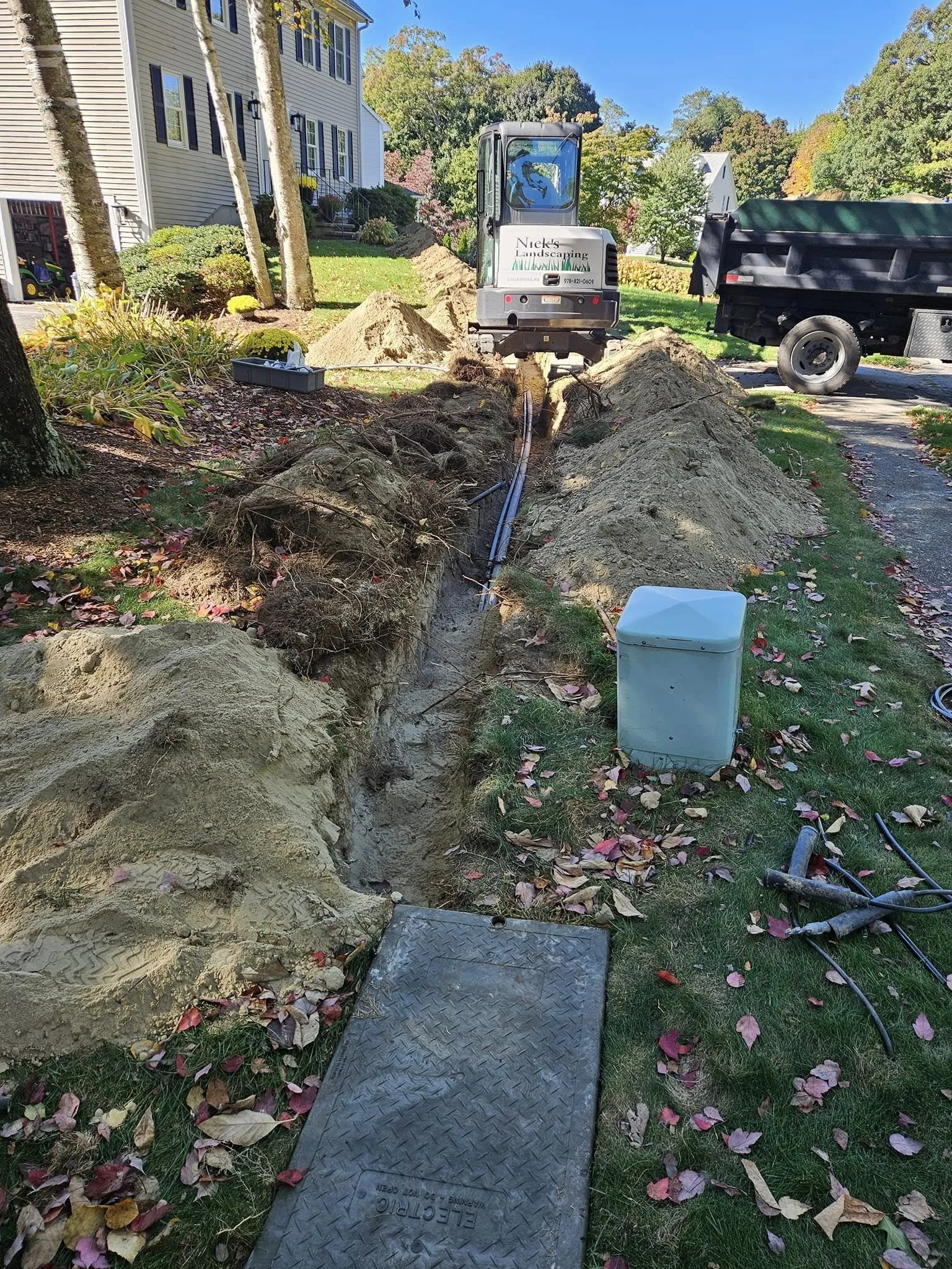 Excavation trench in a residential yard surrounded by grass and fallen autumn leaves, with construction equipment and tools nearby, including a small excavator and a truck, under a clear blue sky.