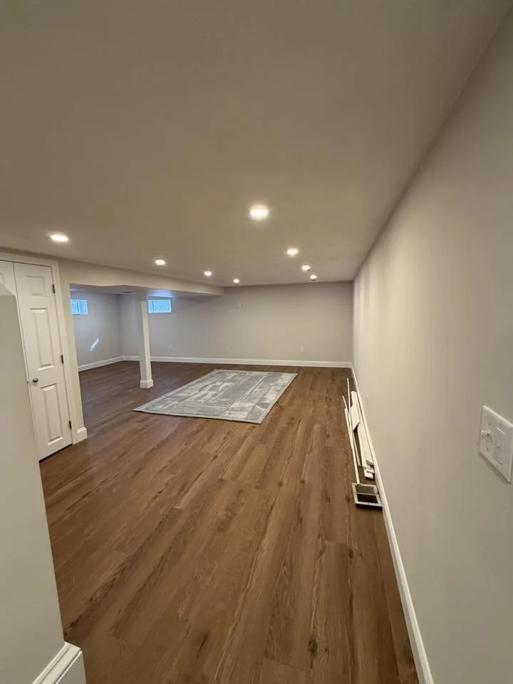 Empty basement room with hardwood floors, recessed ceiling lights, small windows, a closed door, a light-colored area rug, and some baseboards or heating units on the right side.