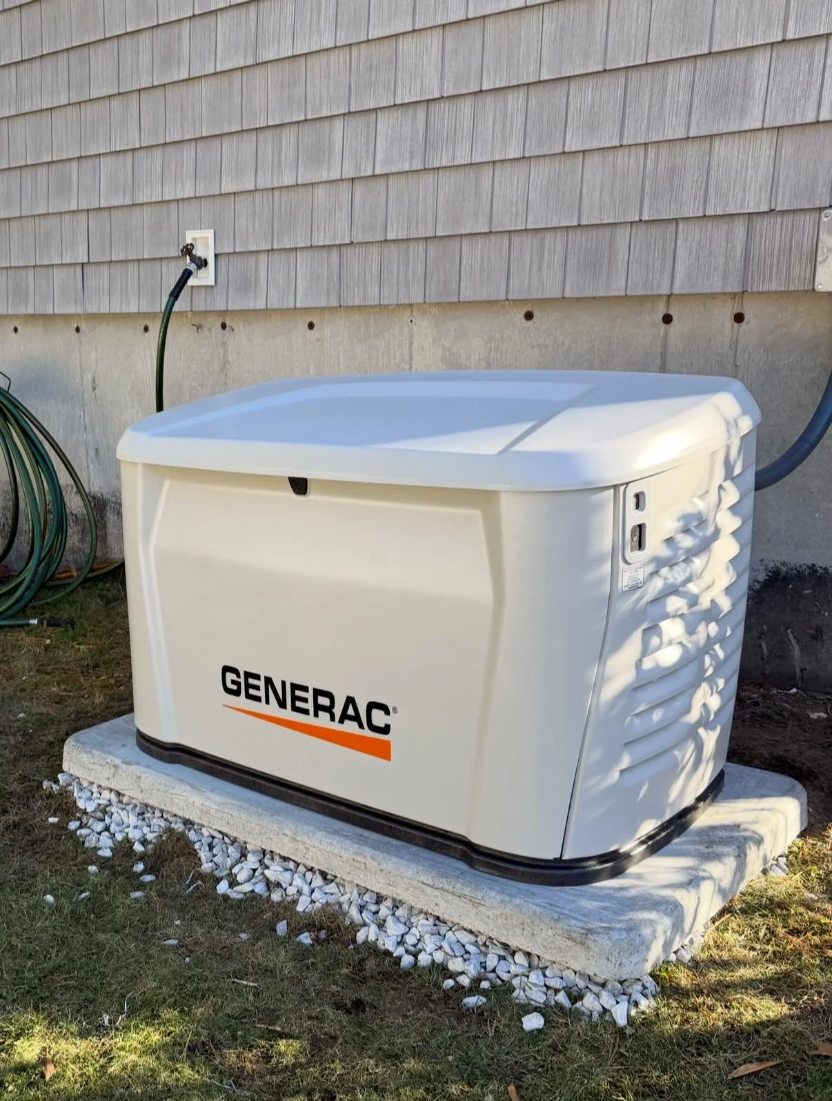 Photo of a Generac generator installed outside a house on a concrete slab, with a garden hose hanging on the wall behind it.