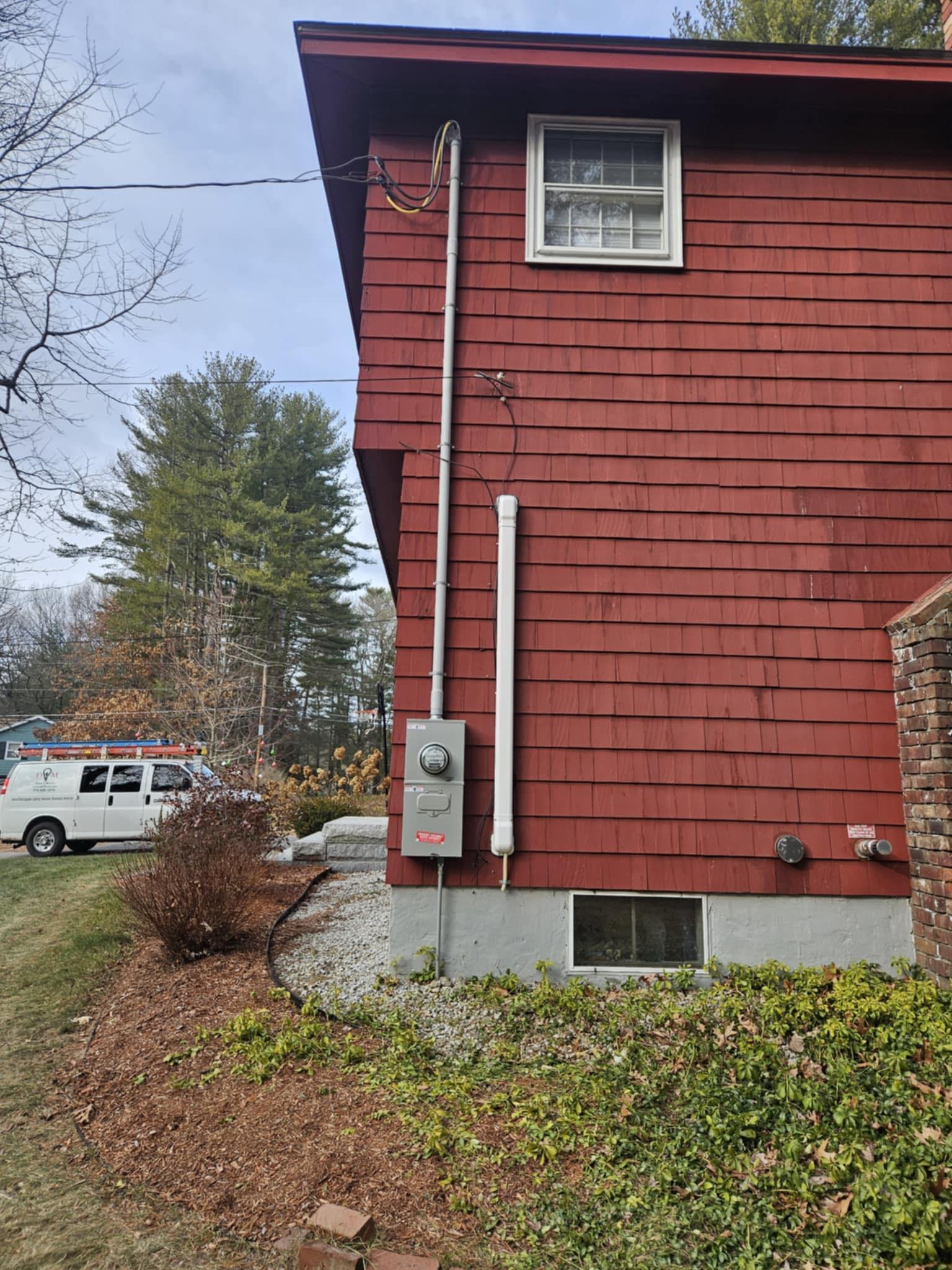 The side of a red house with siding, a window, and utility equipment including a meter and electrical box outside. There are leafless trees and a lawn with bushes and mulch in the foreground.