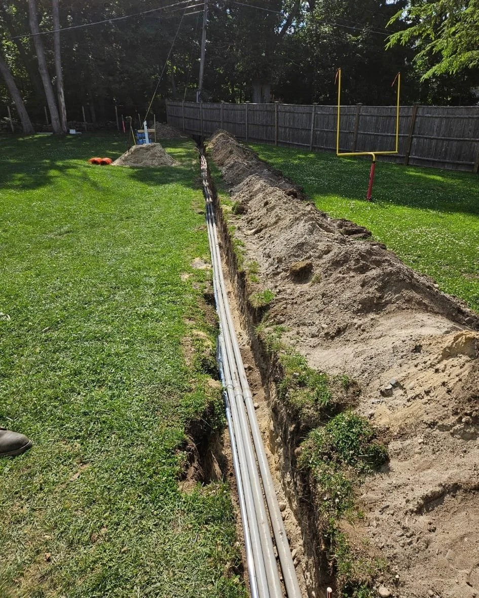 A construction site in a backyard with a long trench dug in the ground, running parallel to a wooden fence. Several metal pipes are laid in the trench, and there's a pile of dirt at the far end. On the grassy area, there's an American football goalpo