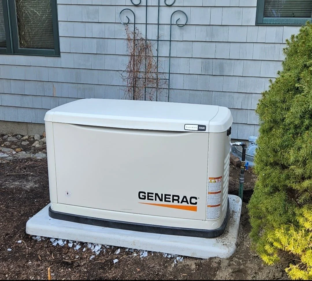 A Generac home standby generator outside a house on a concrete pad, next to a green bush, with a gray house wall in the background.