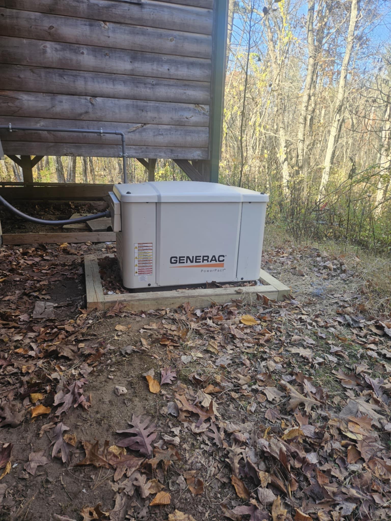 A portable Generac generator placed on a wooden stand outdoors among fallen autumn leaves, with trees and a wooden building in the background on a clear day.