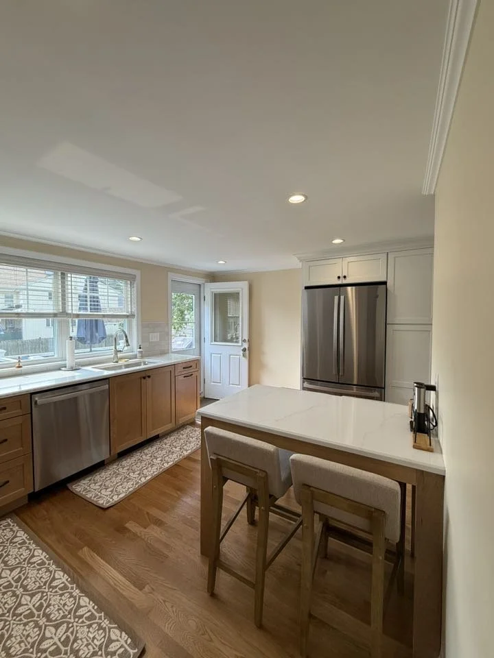 Bright kitchen with wooden cabinets, stainless steel appliances, a white marble island, a window above the sink, and hardwood floors.