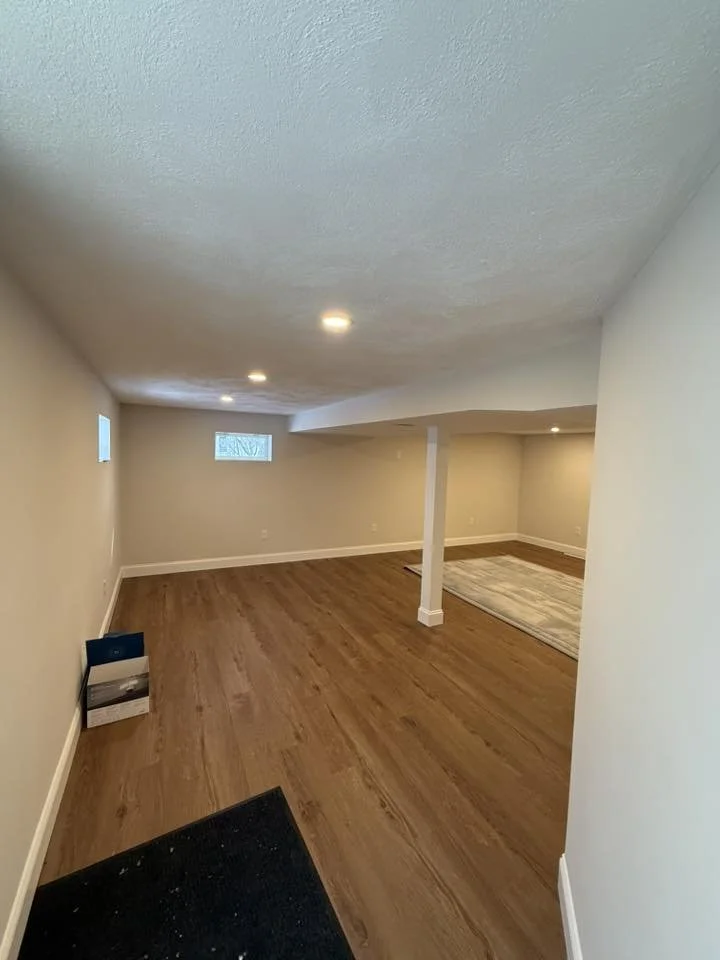 Empty basement with wood flooring, beige walls, small windows, and recessed lighting, divided by a support column, with a small rug and some boxes in the corner.