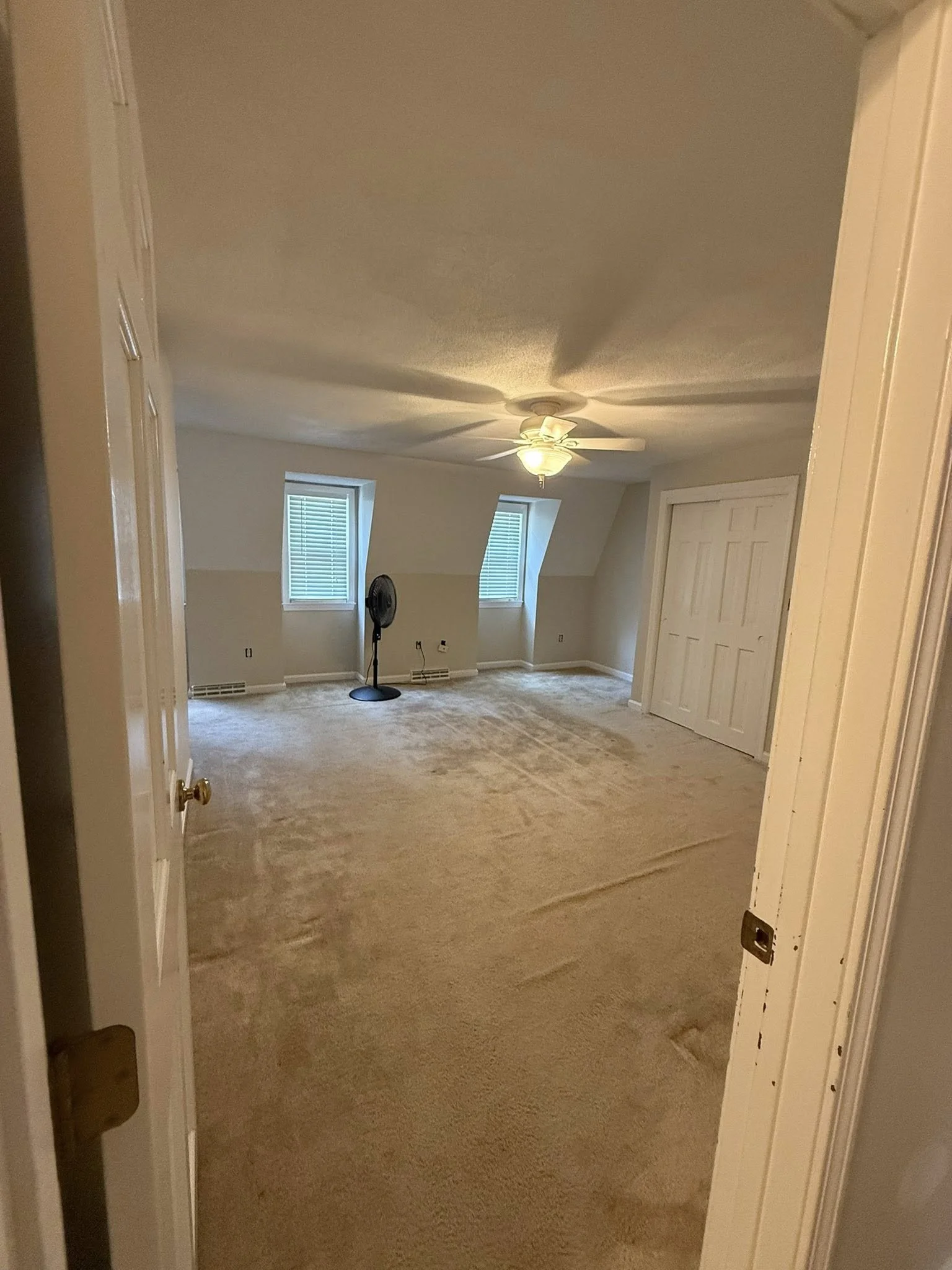 Empty room with beige carpet, two small windows with blinds, a ceiling fan with light, and a black standing fan.