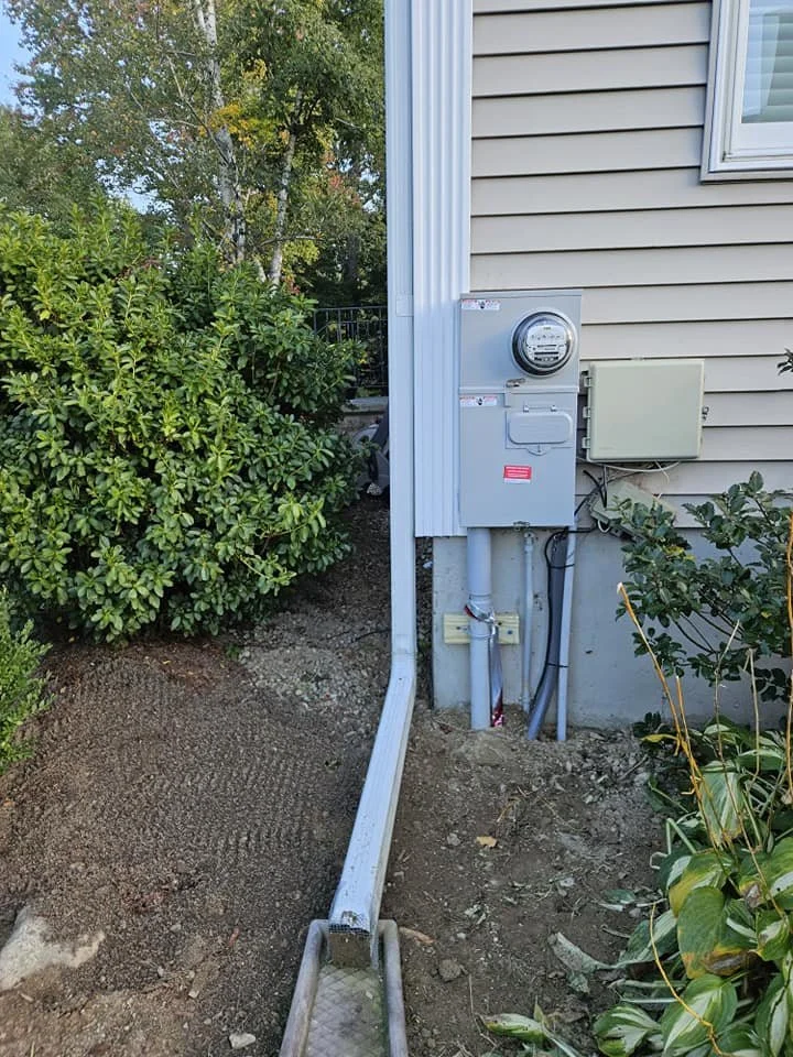 Side of a house with an electrical meter, electrical box, and conduit pipes, next to green bushes and soil.