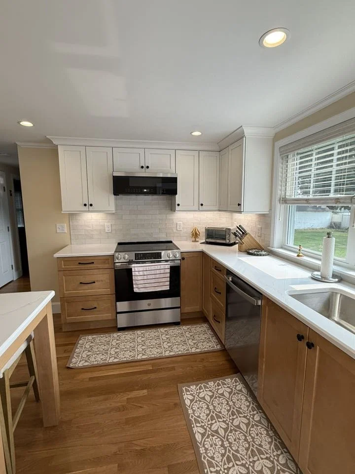 A modern kitchen with white upper cabinets, wooden lower cabinets, and stainless steel appliances including an oven and dishwasher. There is a window with blinds, a white countertop, and a white subway tile backsplash. The floor is wooden, and there 