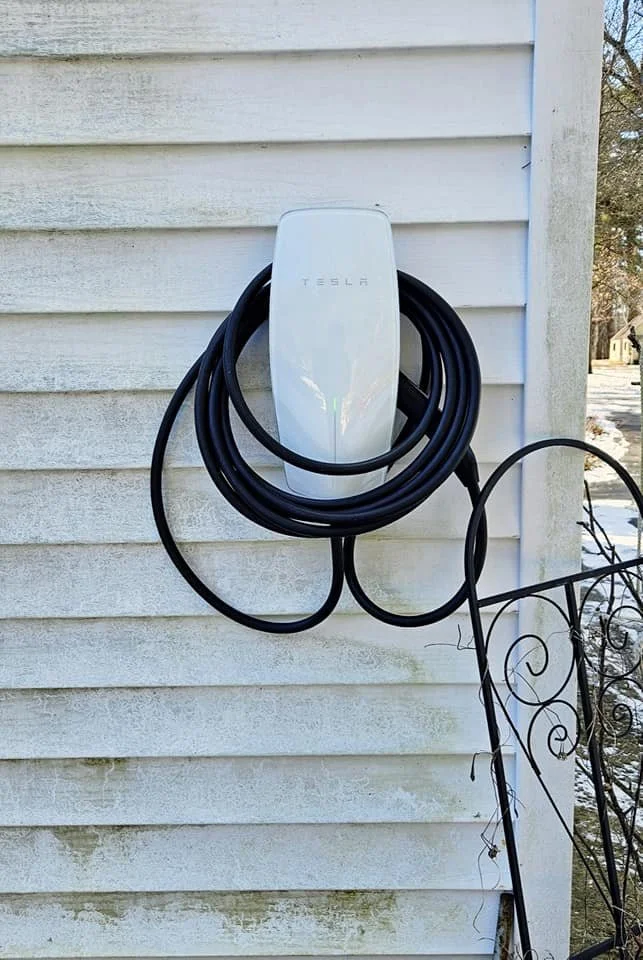 Tesla electric vehicle charging station mounted on a house's white siding with a coiled charging cable hanging on it.