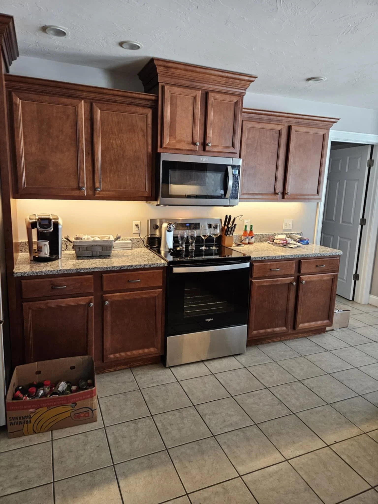 A kitchen with wooden cabinets, stainless steel microwave and oven, granite countertop, coffee maker, and a box of spices on the floor.