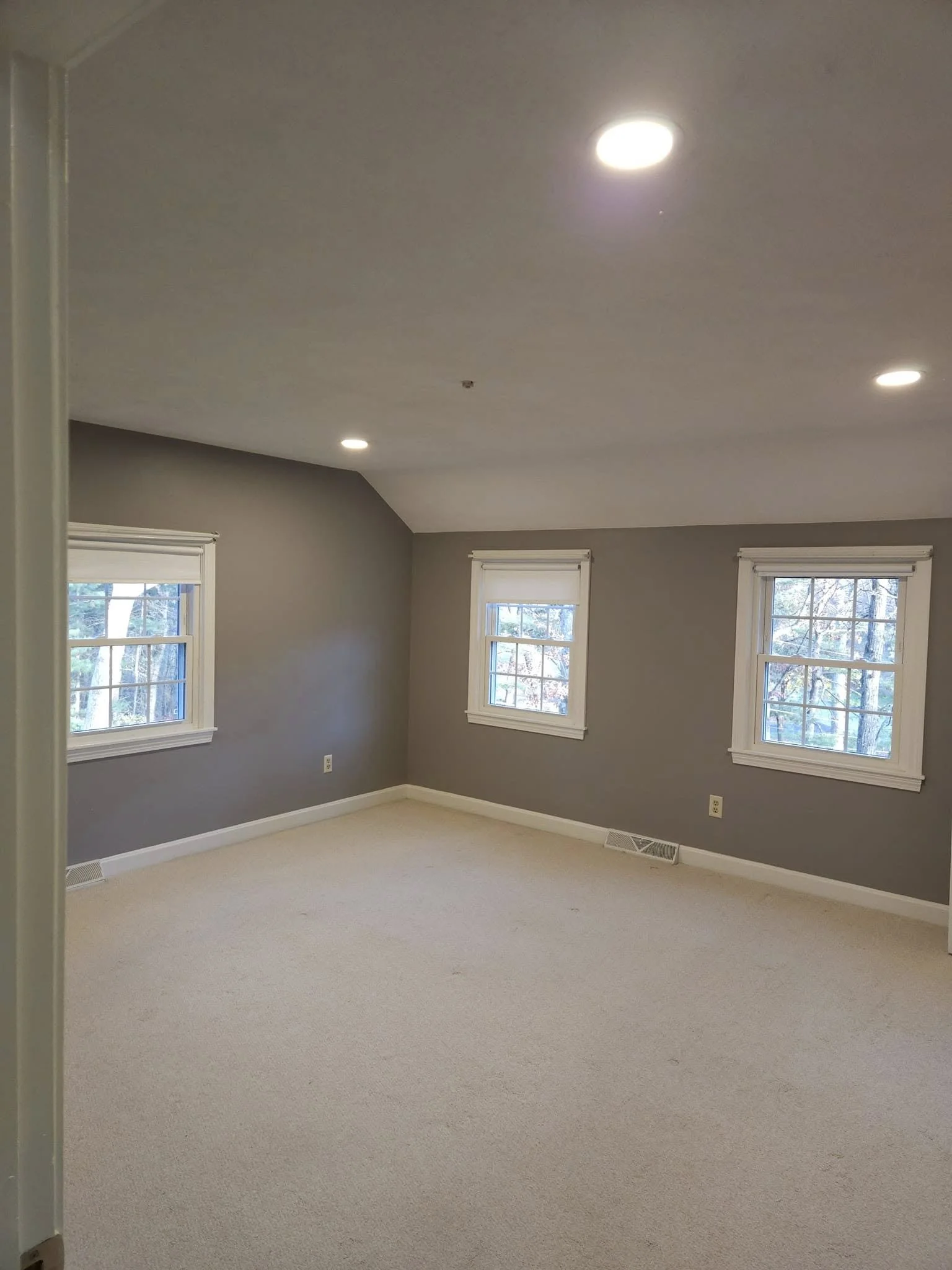 Empty room with beige carpet, gray walls, three windows with white frames, and recessed ceiling lights.