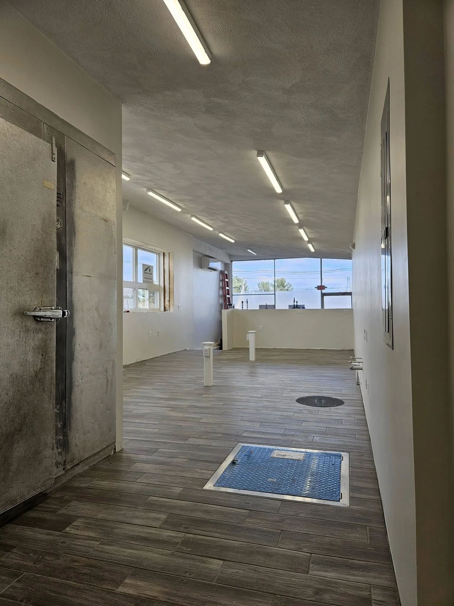 Interior of a modern commercial space with wooden flooring, white walls, fluorescent lighting, large windows, and an emergency exit door.