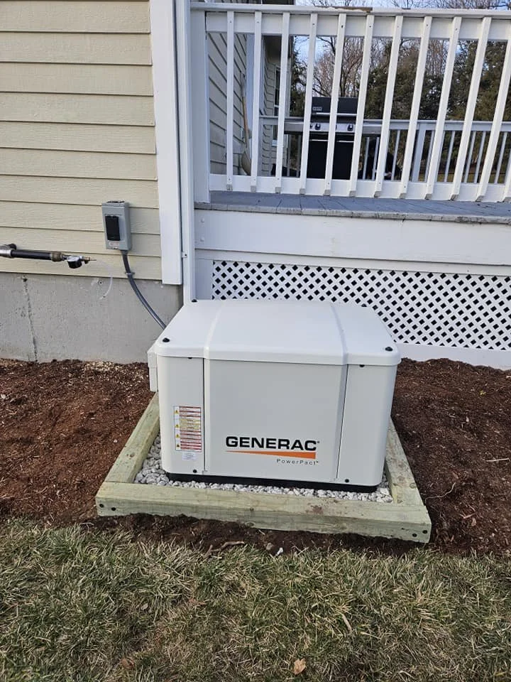 A Generac PowerPact portable generator installed on a wooden platform outside a house with beige siding, next to a small patch of grass and soil.