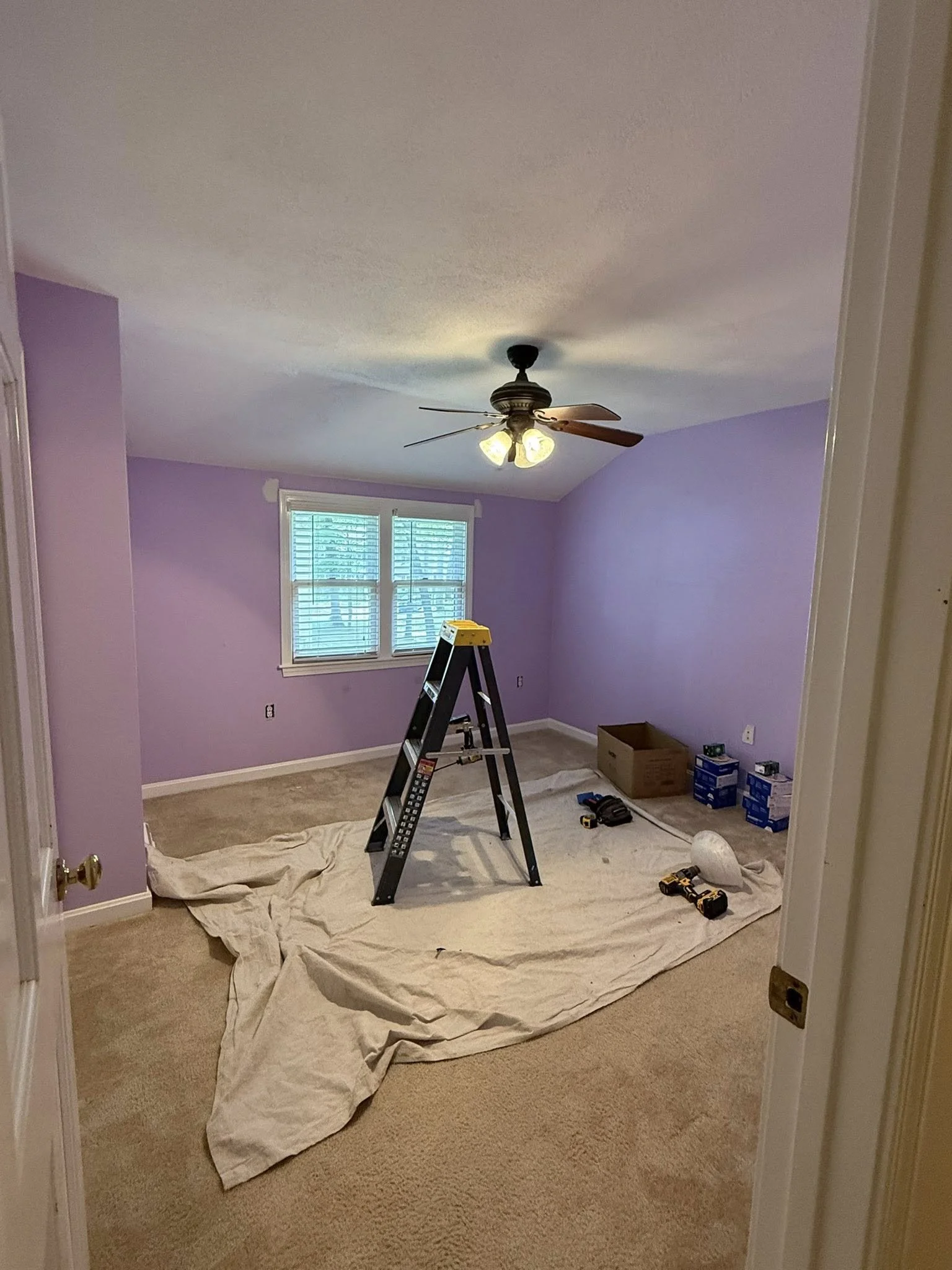 A room with lavender-colored walls and beige carpet, under renovation. A stepladder, tools, and boxes are scattered on the floor, with a drop cloth underneath. The ceiling fan has four lights and wooden blades, and the room has a window with blinds.