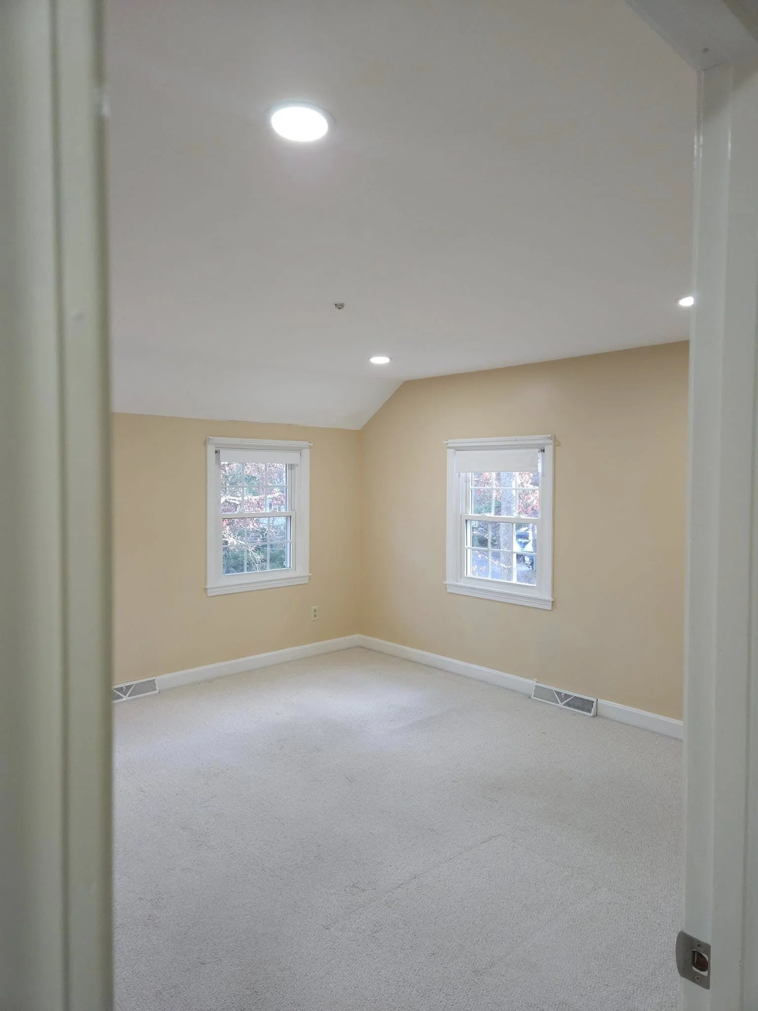 A room with cream-colored walls, two windows with white frames, a beige carpet, and ceiling lights, viewed through a doorway.