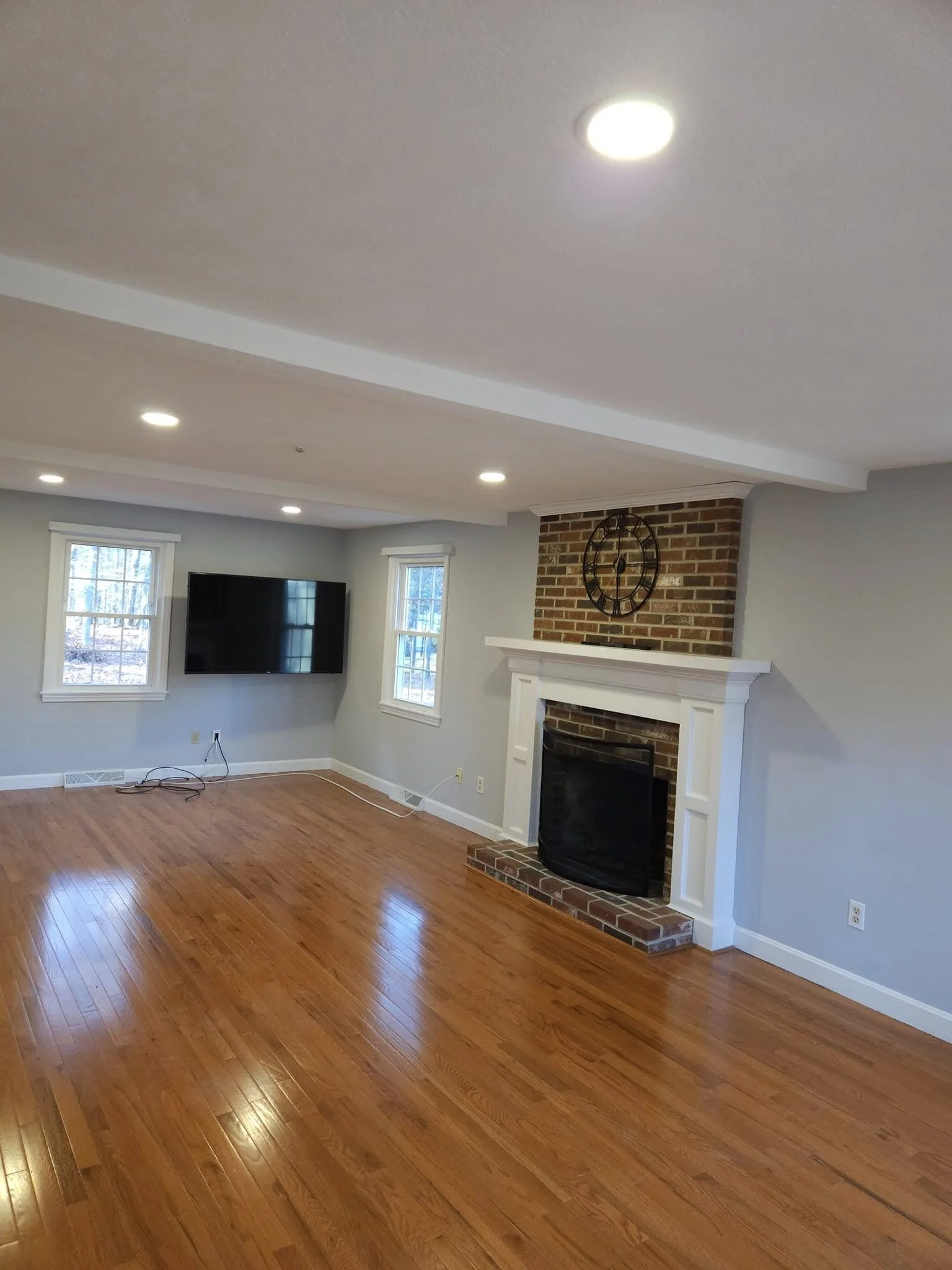 Living room with hardwood floors, a brick fireplace with a white mantel, a large wall clock, a flat-screen TV, and two windows letting in natural light.