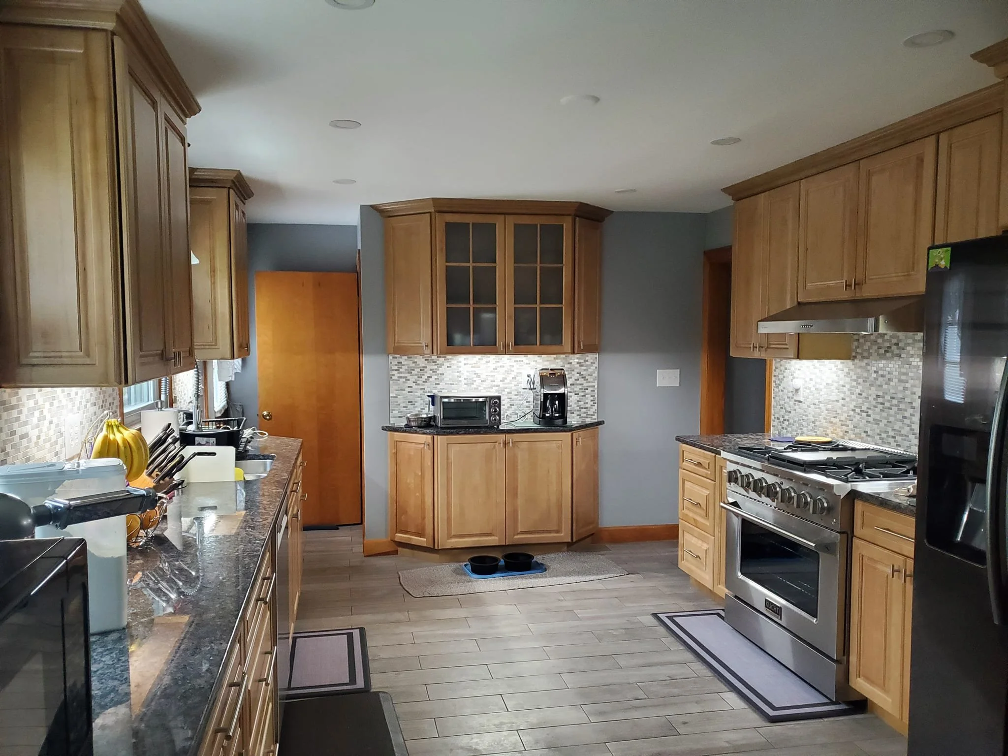 Kitchen with wooden cabinets, stainless steel stove, microwave, toaster oven, coffee maker, and a counter with bananas and other items. Tiled backsplash and hardwood flooring.