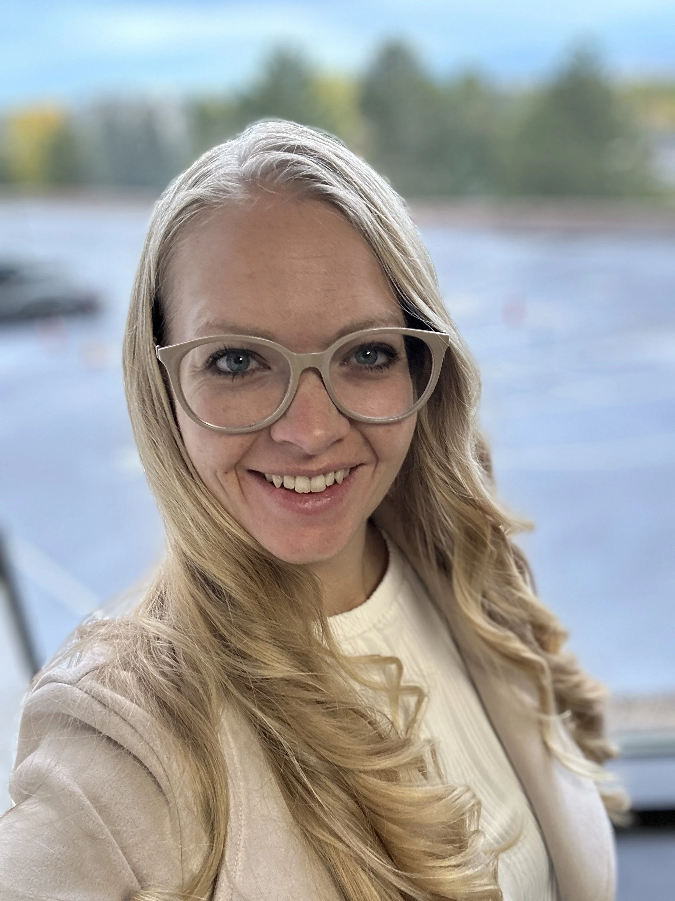 A woman with long blonde hair, wearing glasses, smiling, in an indoor setting with a blurred blue and green background.