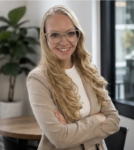 A smiling woman with long blonde hair, glasses, and a beige blazer standing in an office.