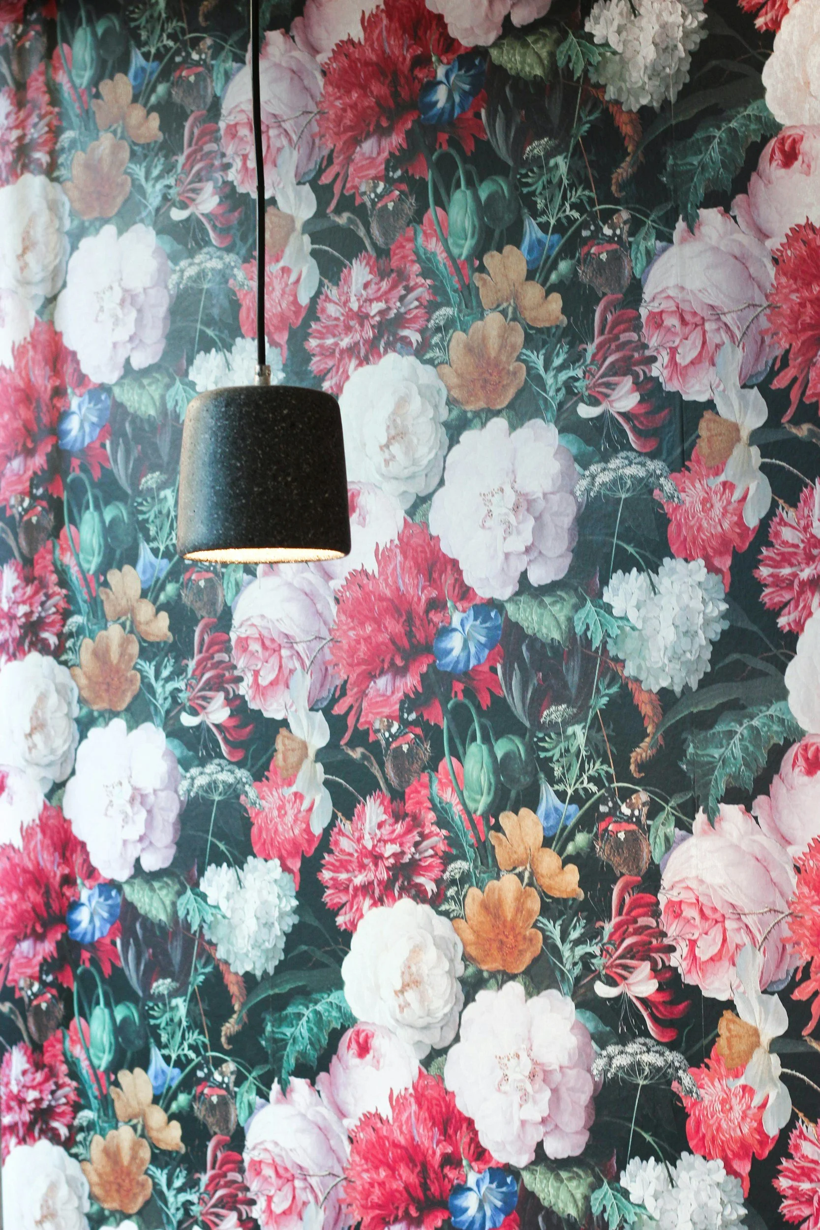 A wall covered in floral wallpaper with large pink, white, red, blue, and cream flowers and green leaves, with a black hanging pendant light.