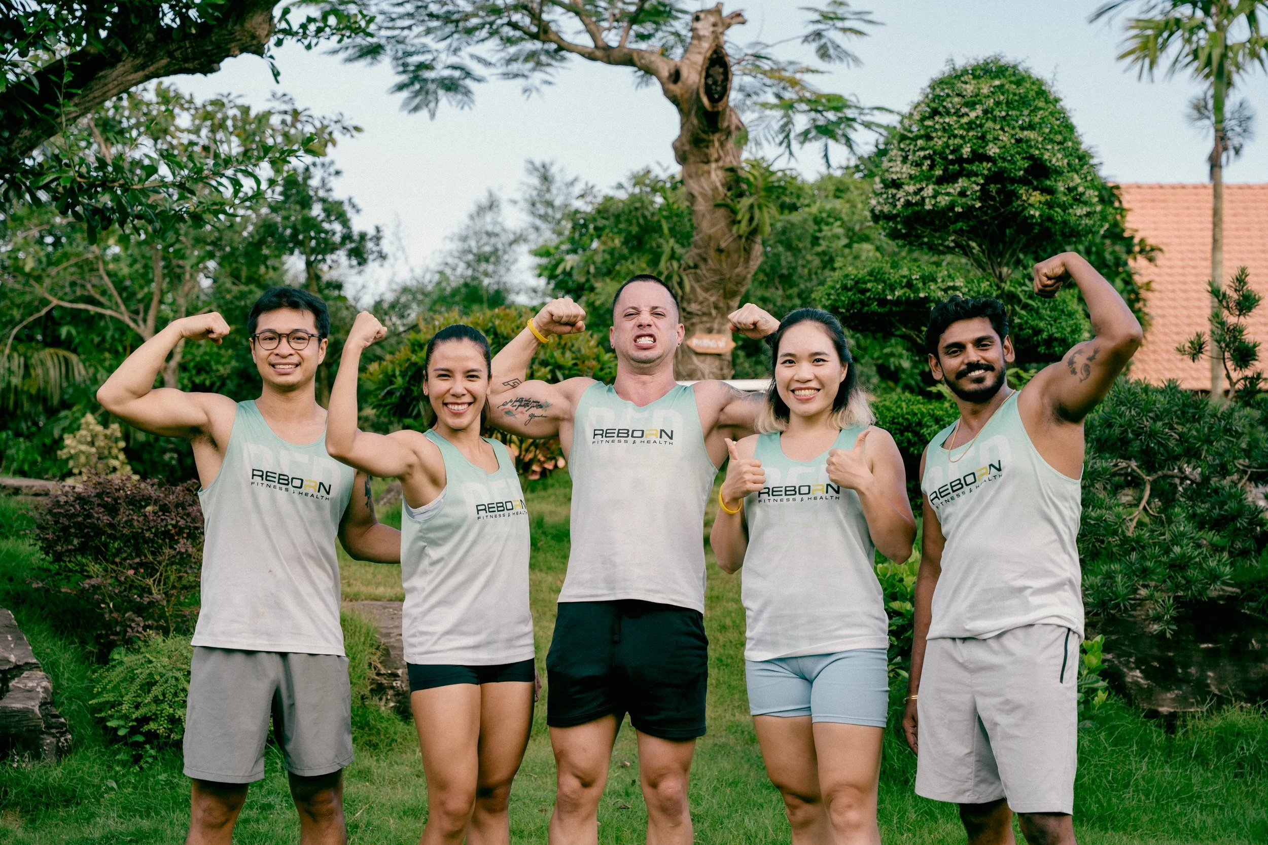 Group of five diverse young adults standing outdoors in a park with trees and greenery, flexing their arm muscles and smiling.