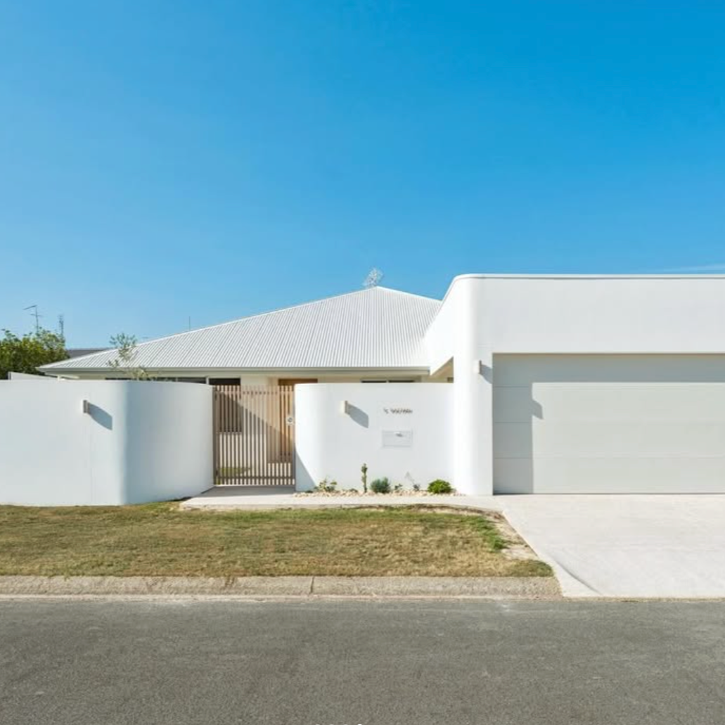 Modern white house with a metal roof, white curved wall, and a gated entrance, with a driveway, small front garden, and a clear blue sky.