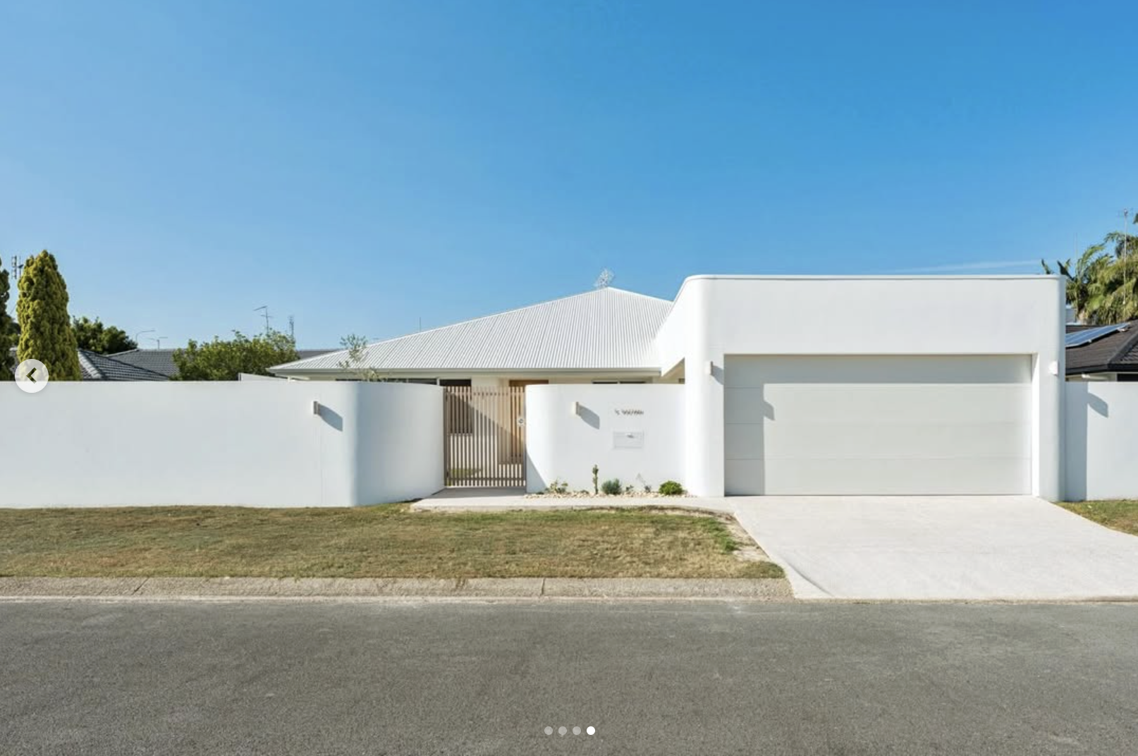 Modern white house with a flat roof and a tall white wall, a wooden gate, and a two-car garage. Minimal landscaping with a small patch of grass and some plants in front.