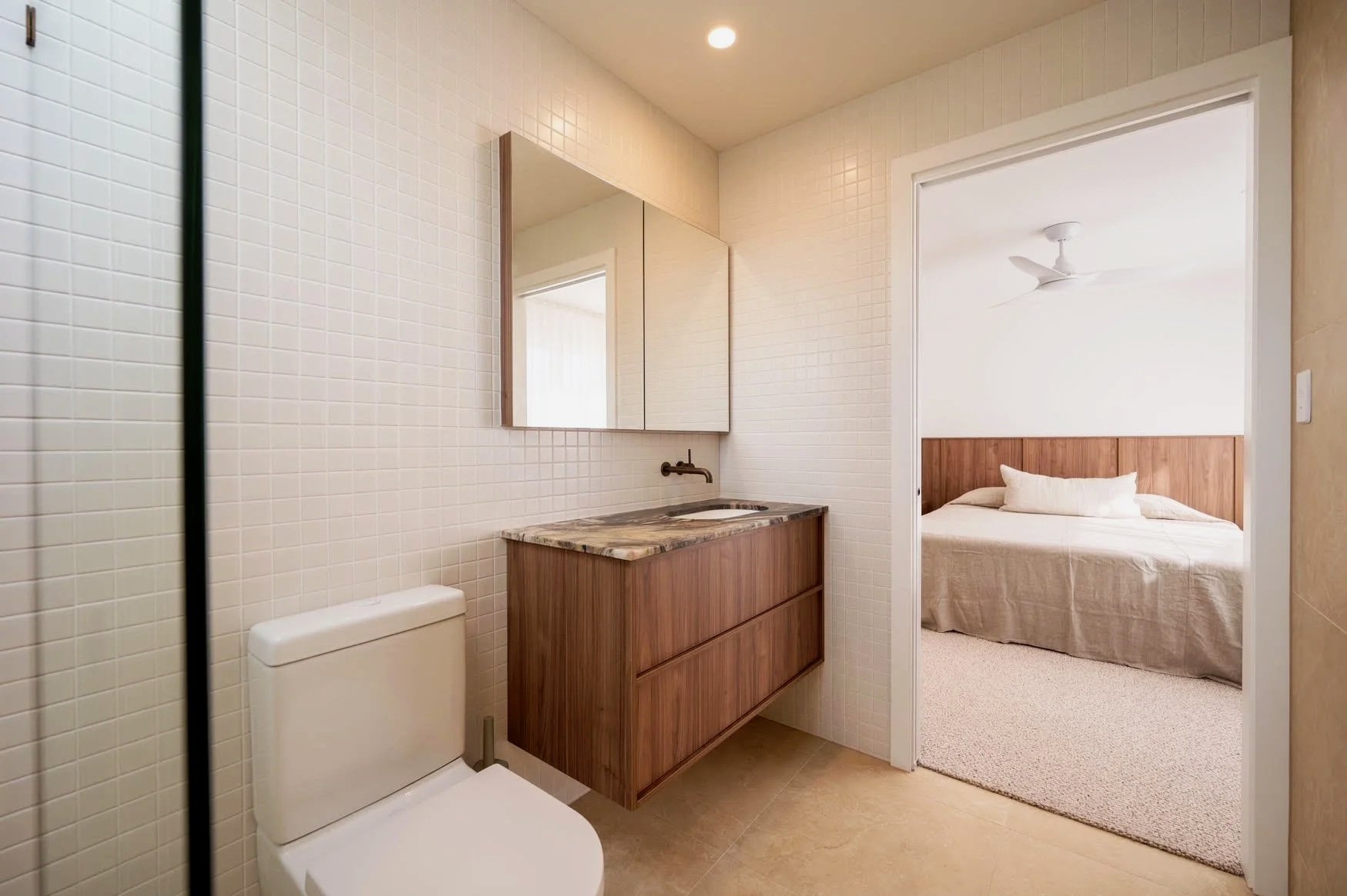 Bathroom with beige tiled walls, wooden vanity with marble countertop, mirror, toilet, adjacent bedroom with bed, nightstand, and ceiling fan.